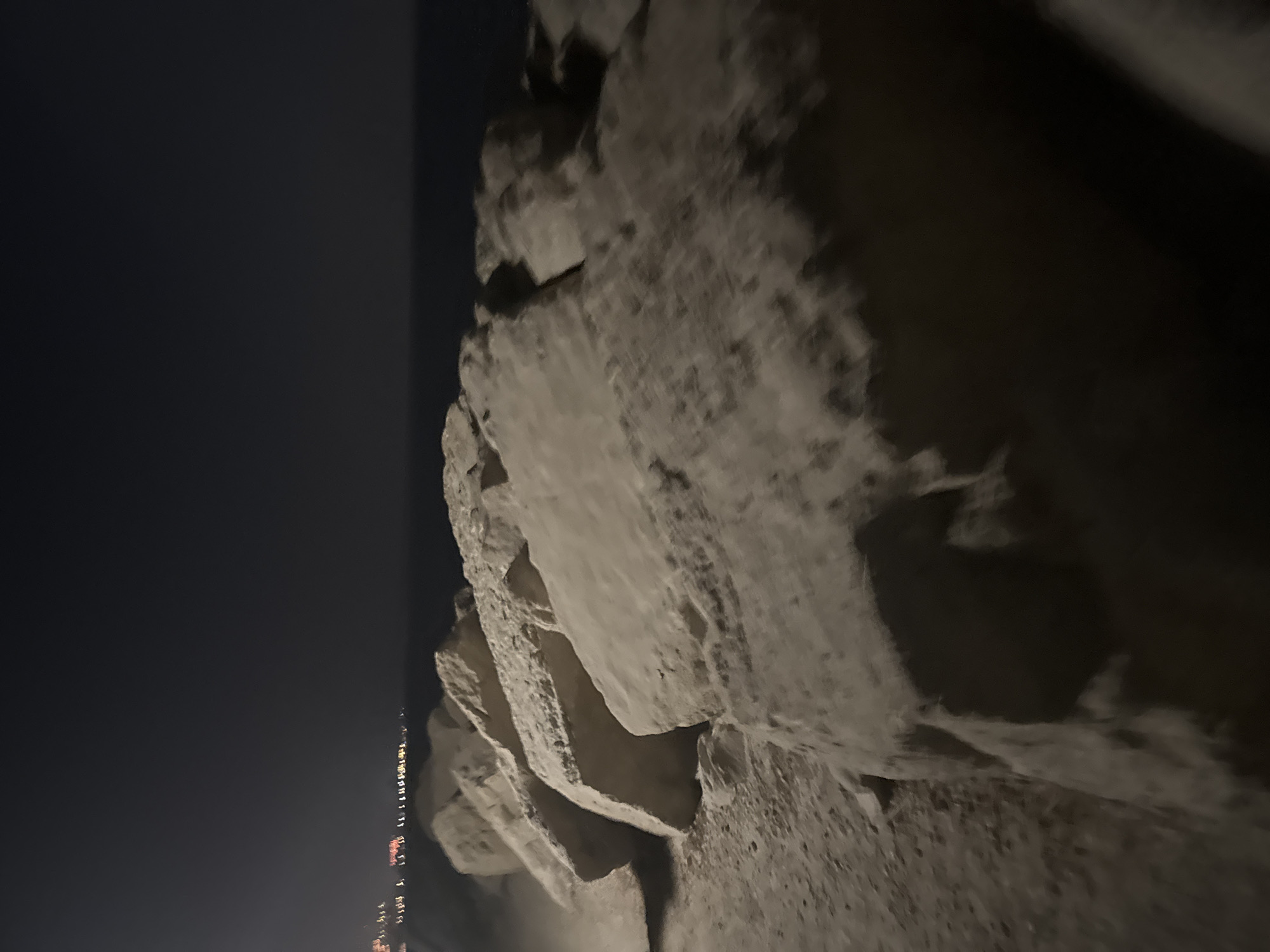 The image captures a nighttime scene of a rocky shoreline with a distant view of city lights. Large, rough-hewn rocks dominate the foreground, arranged in a diagonal line receding into the distance. The rocks are a pale, sandy color and show texture and natural imperfections. The ground beneath the rocks appears to be sand or gravel.
In the background, the dark expanse of the sea meets a hazy horizon. Faint, blurred lights of a city or town are visible along the coastline, suggesting an urban or developed area in the distance. The overall atmosphere is dark and somewhat moody, illuminated primarily by the distant city lights and the ambient darkness.
The location is identified as the Agios Tychona Community in Cyprus. The scene likely depicts a breakwater, pier, or natural rocky formation along the coast of Agios Tychona at night. There are no visible people or active interactions depicted in the image. The focus is on the geological formations and the distant urban illumination.