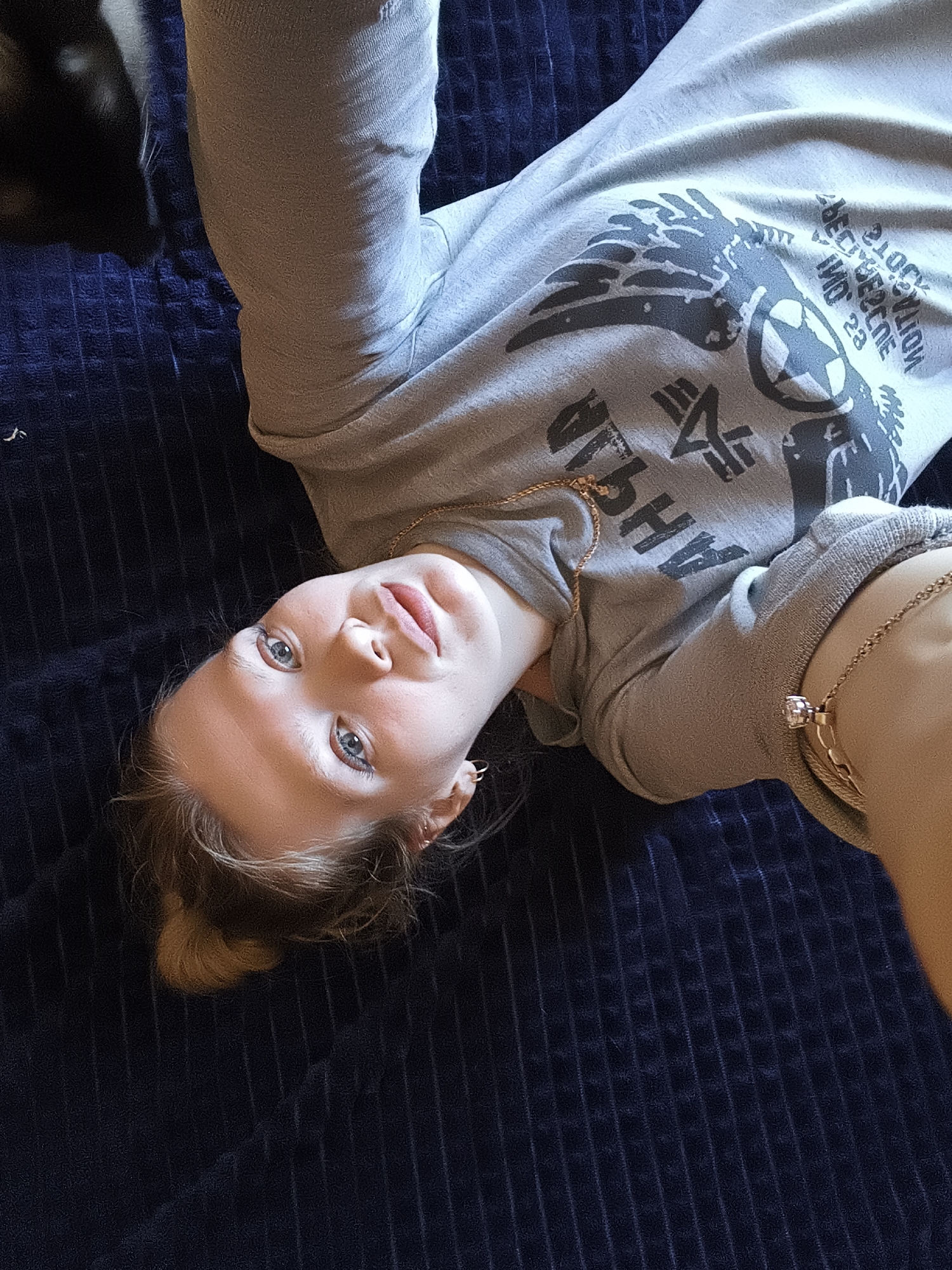A young woman with light hair and blue eyes is lying on a dark blue corduroy surface, possibly a bed or couch. She is wearing a light gray sweatshirt with a graphic design including the word "ALPHA" and a star-like symbol. She has jewelry on, including a necklace and a bracelet with a large stone. Part of a dark object, possibly a pet, is visible in the upper left corner. The image appears to be taken from a slightly overhead, selfie perspective. There is no indication of Русский Вожой, Russia.