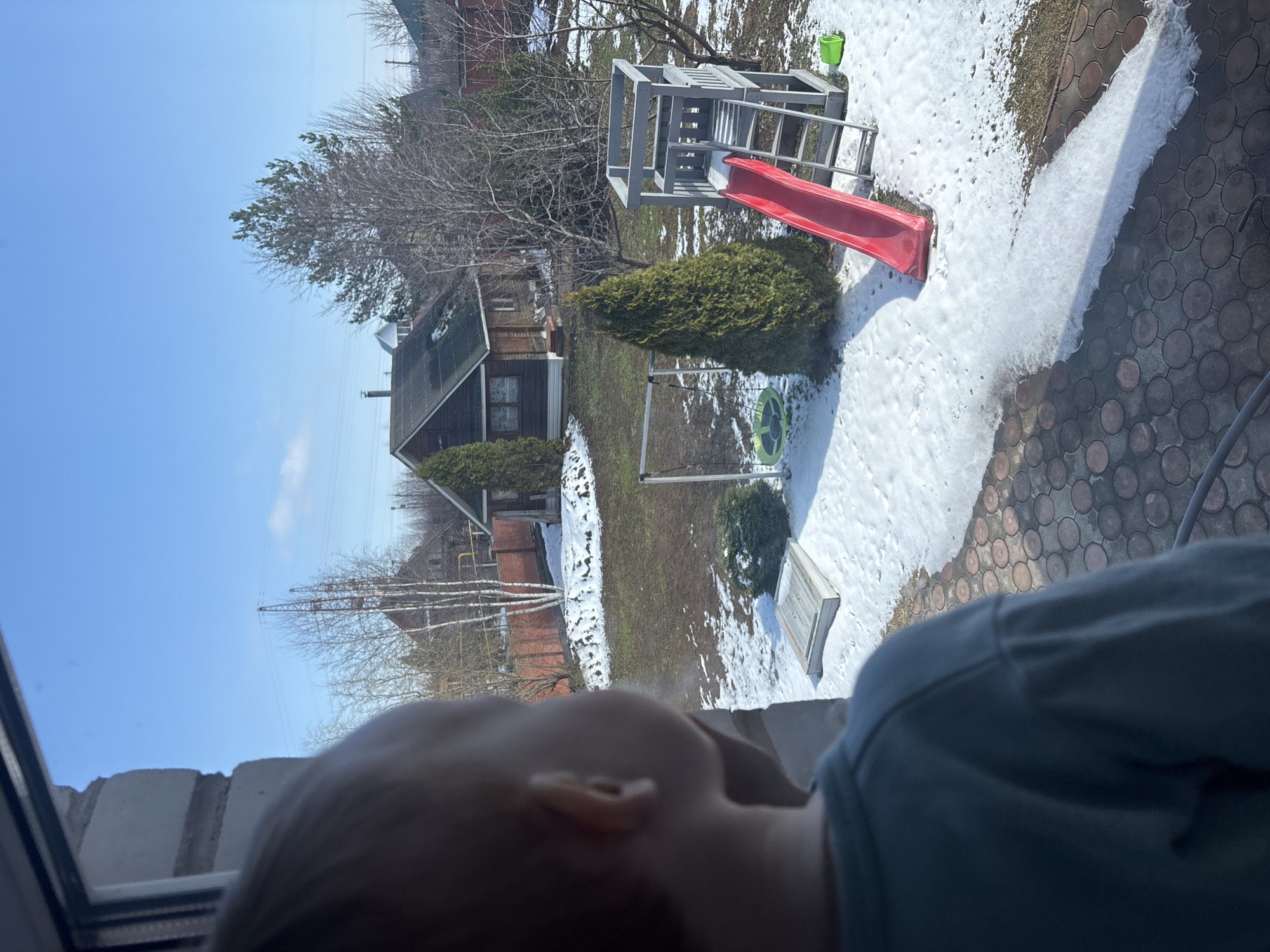 A partially visible child looks out a window at a backyard in Ferma, Russia. Patches of snow cover a green lawn, and a wooden play structure with a red slide sits near a green bucket. A small cabin-like house with a dark roof and brown siding is visible in the background. Bare trees stand next to the house. A brick-patterned ground surface extends from the house, partially covered by snow. A power line runs across the blue sky.