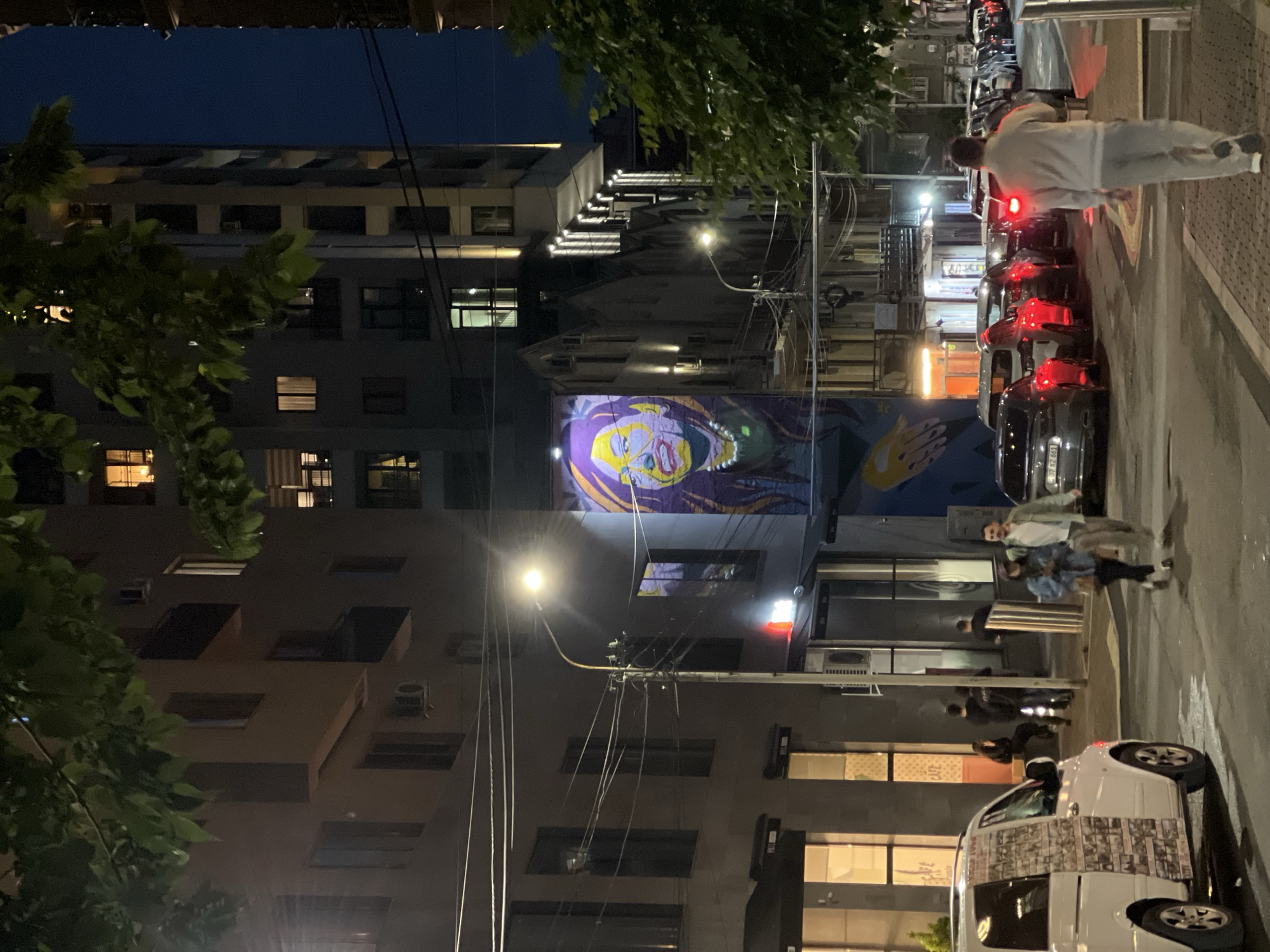 At night in Yerevan, Armenia, a street scene reveals parked cars, pedestrians, and buildings. A large mural with a face is visible on a building, and power lines crisscross above. People are walking on the sidewalk, and cars are lined up on the street. The lighting is provided by streetlights and building lights. The overall atmosphere is urban and active.