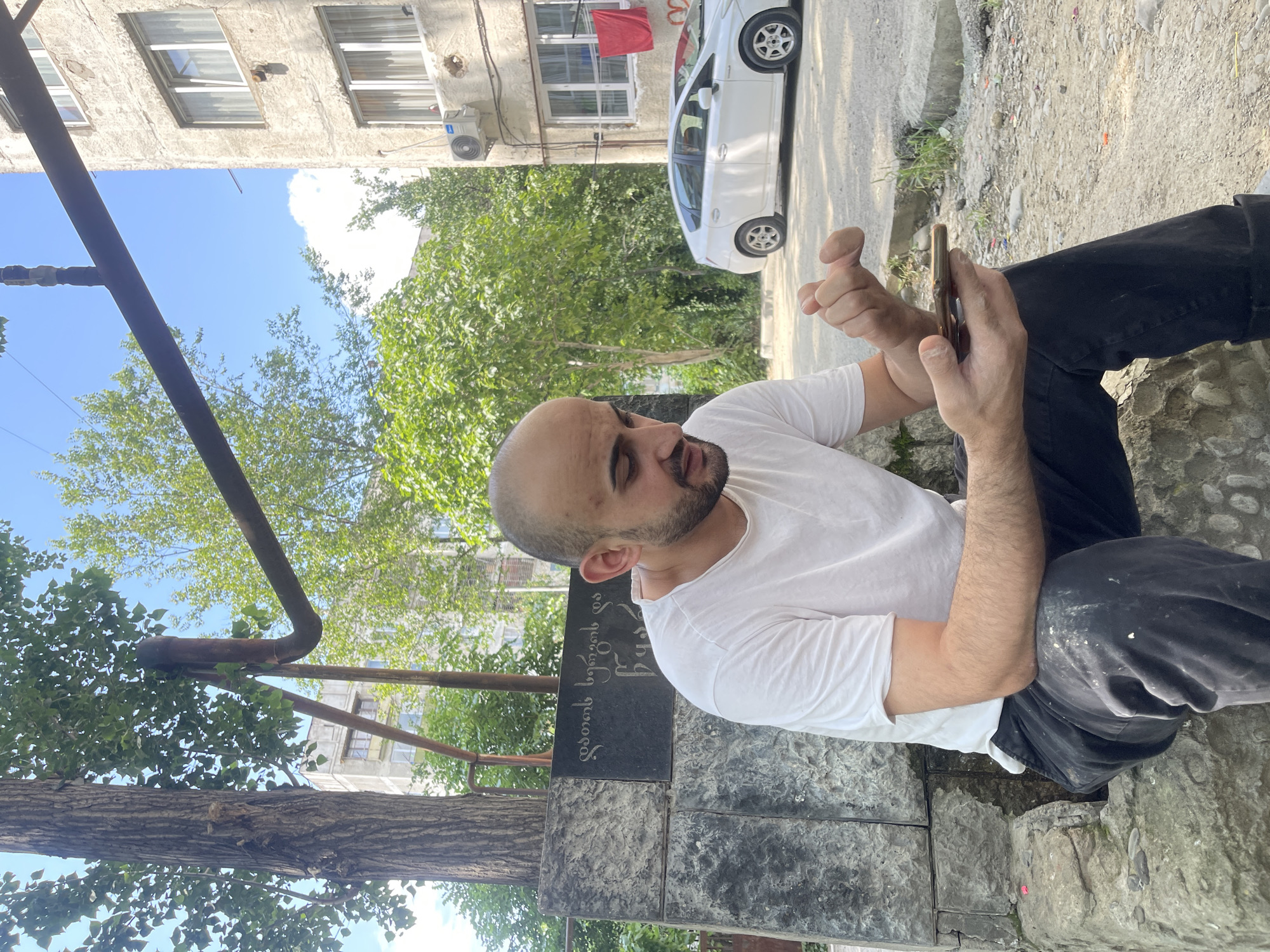 A bald, bearded man with a light complexion is sitting on a stone structure outdoors, looking at his mobile phone. He is wearing a white t-shirt and black pants. To his left, there is a stone monument with writing in Georgian script. Behind him, a large tree stands to the left, and an apartment building is visible to the right. A white car is parked in front of the building. The scene appears to be in a residential area of Tbilisi, Georgia. The man's posture and focus on his phone suggest he is engaged with its content. The overall environment indicates a casual, outdoor setting, possibly a courtyard or public space.