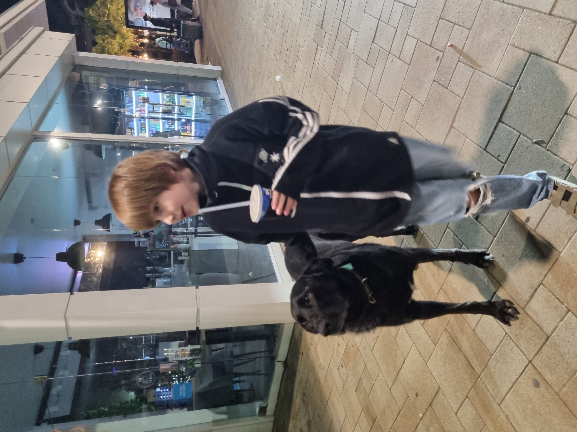 A young person stands outside a building with a large window in Sochi, Russia. The person, with short brown hair, wears a dark jacket with white stripes, ripped jeans, and sneakers, and holds a cup. A black dog sits beside the person. The building has a brightly lit interior visible through the window, showing shelves of drinks. The ground is paved with rectangular blocks.
