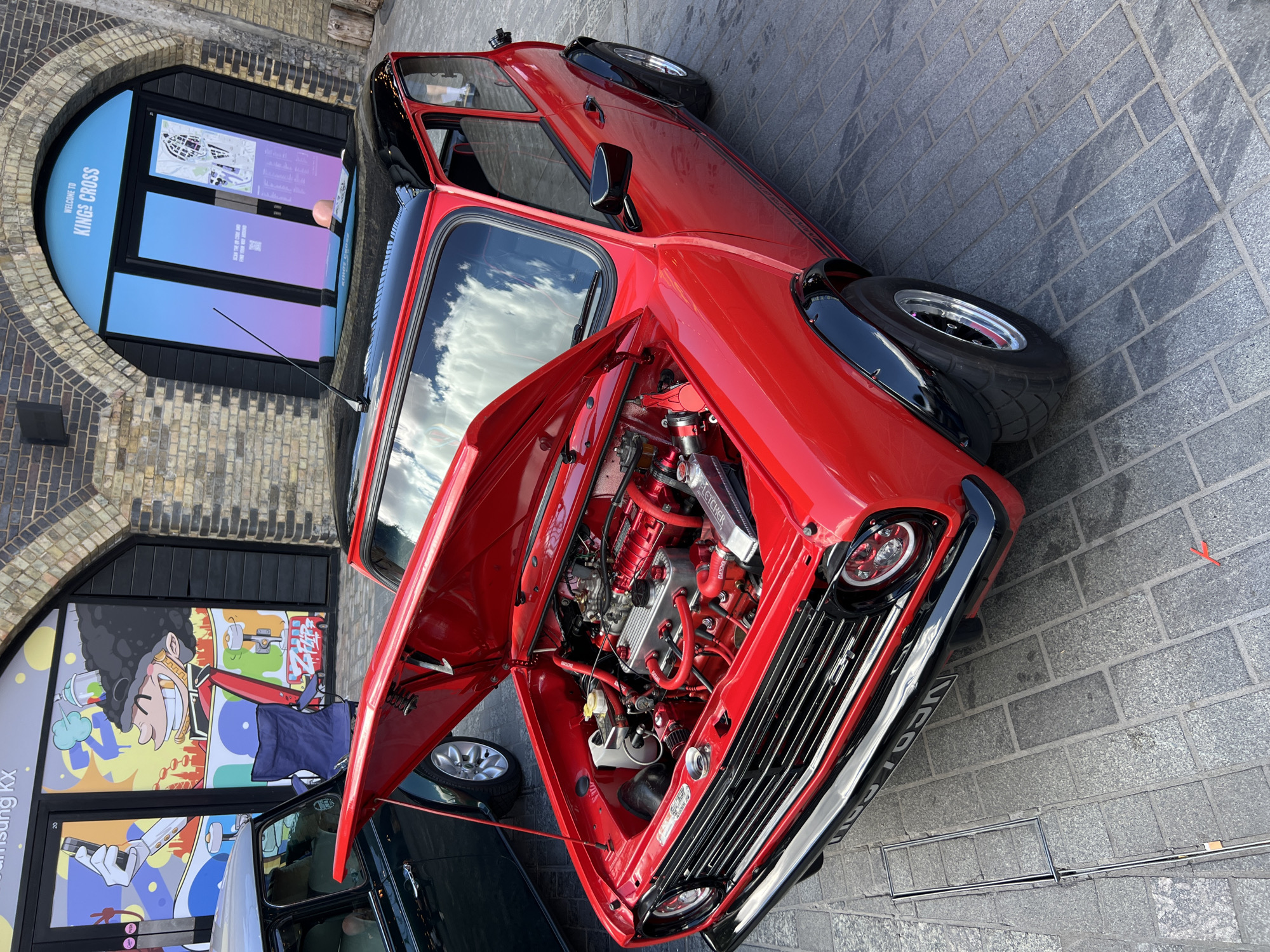 The image presents a vibrant red classic Mini car parked on a grey paved surface in what appears to be an urban setting in London, United Kingdom. The car's bonnet is open, revealing a highly customized engine bay with many components, including hoses and covers, painted in a matching bright red, contrasted with silver engine parts. The text "FLETCHER" is visible on a radiator or intercooler, and "ROCKER" on a hose clip. The front of the Mini features a prominent chrome grille, round headlights, and black fender flares over wide tires on alloy wheels. A partially visible license plate reads "VE02 C...N".
To the left, a portion of another dark-colored classic Mini is visible, suggesting a gathering or exhibition of these vehicles. The background features a brick building with large arched and rectangular window displays. On an arched window, white text clearly states "WELCOME TO KINGS CROSS" and "Scan the QR code and explore the district," alongside a stylized map. Further left, a separate window display shows colorful graphic art and the text "Samsung KX" and "DO NOT TOUCH."
The scene is illuminated by bright daylight, with reflections of a cloudy sky visible in the car's windows, indicating clear weather. The combination of the classic cars, the detailed engine, and the identifiable location text confirms the setting as King's Cross, possibly at an event or display associated with Samsung KX. The meticulous condition of the red Mini suggests it is a show car.