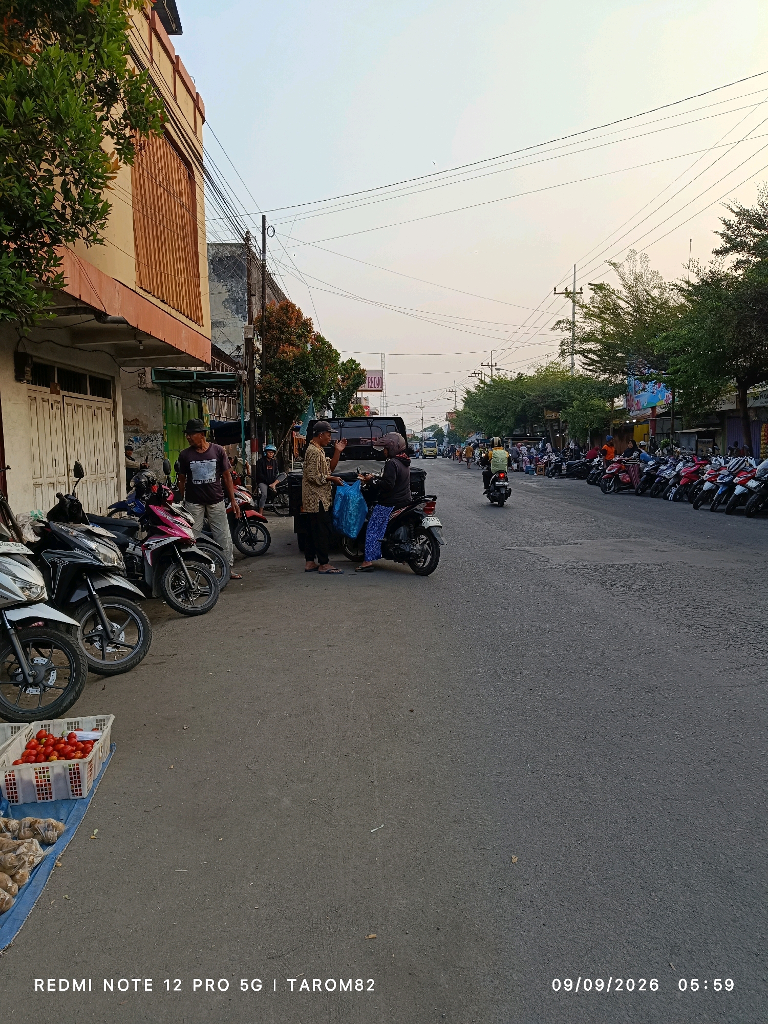 The image captures a street scene in Ngoro, Indonesia, likely a market area based on the presence of vendors and produce. In the foreground, a white plastic basket filled with red tomatoes is visible on a blue tarp, alongside bags of other goods. Several motorcycles are parked along the roadside and sidewalk. A few men are gathered near a parked van, engaged in what appears to be a transaction or conversation, one of them holding a blue plastic bag. A black motorcycle with a rider wearing a helmet is positioned further down the street. The background shows a row of buildings lining the street, with trees and utility poles. The road is paved and appears to be a busy thoroughfare, with more motorcycles parked on the opposite side. The sky is overcast with a soft, diffused light, suggesting it is early morning or late afternoon. The overall atmosphere is that of an active local marketplace.