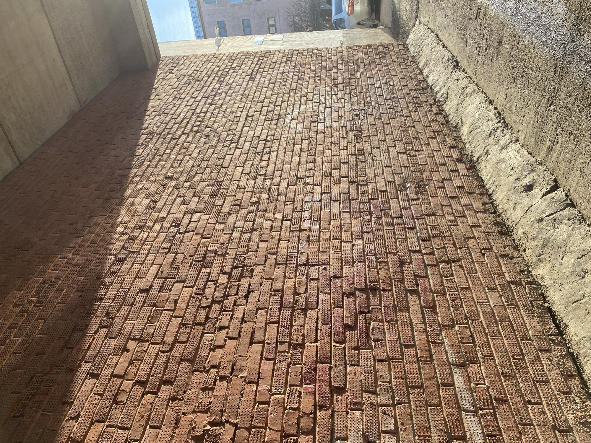 The image depicts a brick street, likely a pedestrian walkway, under an overpass or bridge. The bricks are red and arranged in a running bond pattern. They have a textured surface with small holes. The light suggests the photo was taken during the day. On the left, a concrete support structure provides shade. On the right, the street is bordered by a rough concrete wall or curb. In the background, a building with several windows and a car parked on the street are visible. The location is identified as Noviy, Russia, but there is no specific detail in the image to confirm this.
