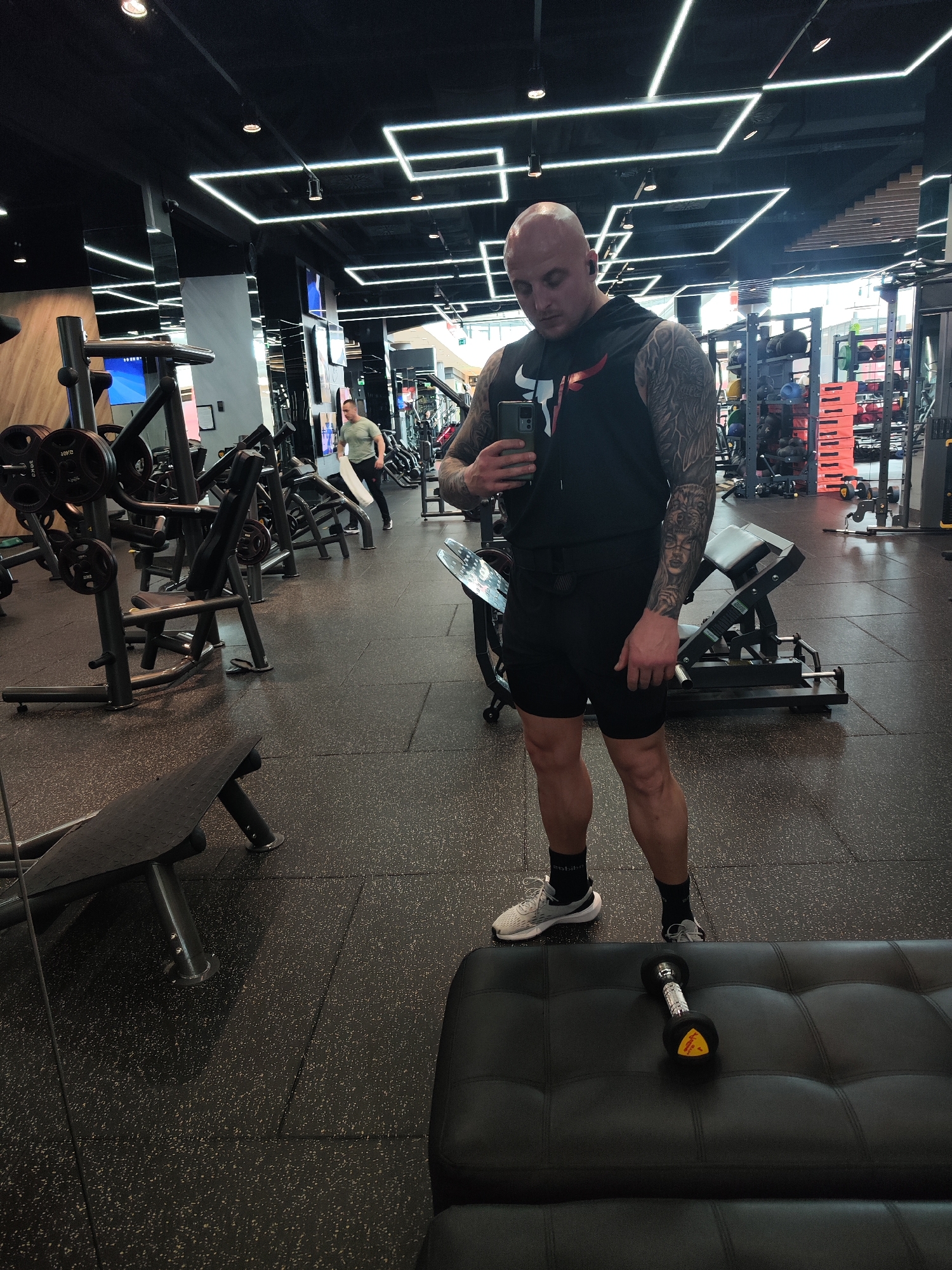 A bald, muscular man with tattoos on his arms is standing in a gym, taking a selfie with his smartphone. He is wearing a black hooded tank top with a bull logo, black shorts, gray athletic shoes, and black socks. A dumbbell is lying on a black upholstered bench nearby. Various weightlifting machines and equipment are visible in the background. Another person is walking in the background, holding a white towel. The gym has a modern design with black floors, mirrored walls, and a grid of white LED lights on the ceiling. The location is likely a gym in Burgas, Bulgaria, as inferred from context and visual cues within the image.