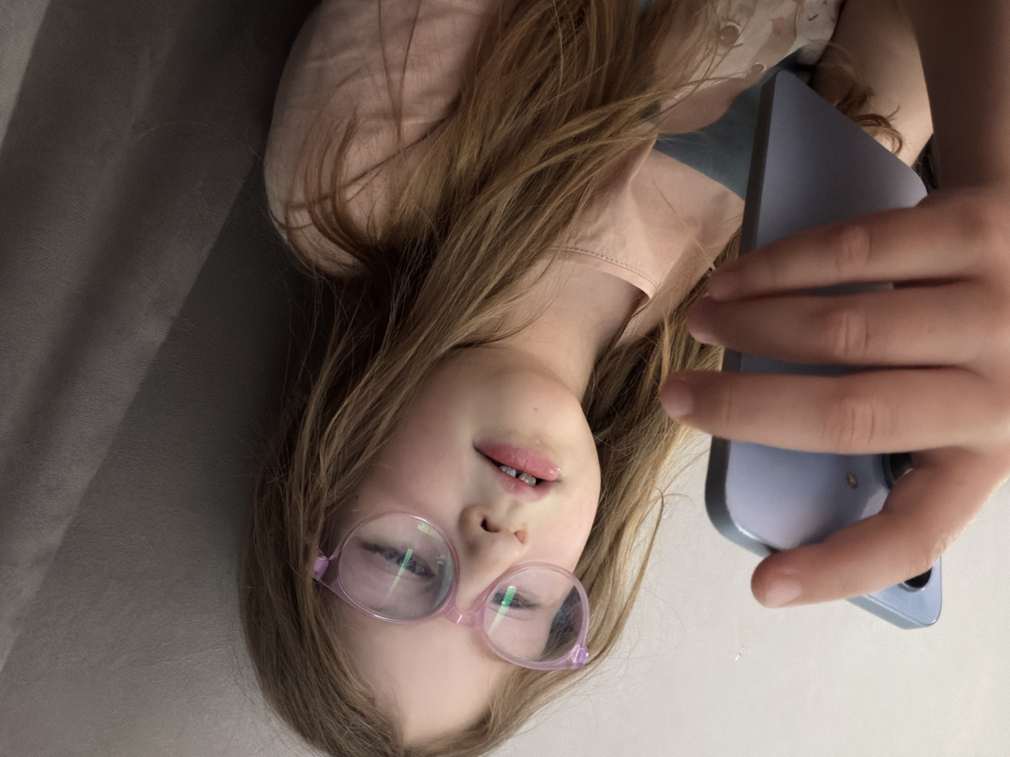 An indoor shot features a young girl lying down, looking directly at a light purple smartphone held in her right hand. The image is oriented with the girl's head to the left and her body extending upwards. She has long, light brown hair spread around her head and shoulders, and is wearing a light pink top. Her face is centered, and she wears round, pink-rimmed glasses, which show reflections of a screen or light source. Her mouth is slightly open, revealing some teeth, and her lips appear glossy. Her expression is relaxed, with a hint of a smile.
The background to her left and behind her head is a dark grey, textured fabric, possibly a sofa or bed. The area to her right and below her is a plain, light, almost white surface, suggesting she is looking up. Her right hand grips the smartphone, with her fingers partially obscuring the device. The activity suggests she is either viewing content on the phone, engaging in a video call, or taking a selfie.
Notable details include the clear reflections in her glasses, indicating direct interaction with a screen, and the overall soft, diffused lighting, typical of an indoor setting. There is no visible text in the image. The location context provided is Chaikovsky, Russia.