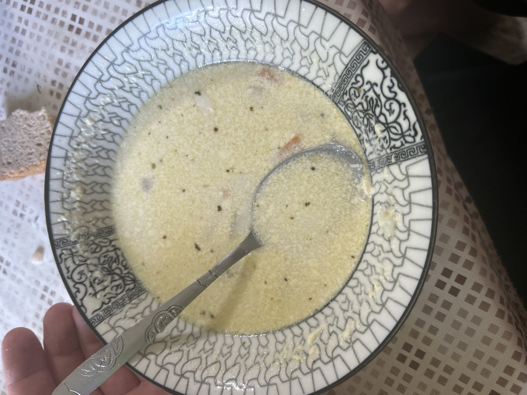 A close-up overhead shot shows a white bowl with a black and white geometric pattern on its rim filled with a creamy yellow soup. A silver spoon is partially submerged in the soup, held by a person's hand which is visible on the left side of the frame. There are some small flecks of green herbs and orange pieces within the soup. A piece of bread is visible on the upper right side of the frame. The setting appears to be a table covered with a patterned tablecloth. The image does not provide any information about the location being in Старотумбагушево, Russia.
