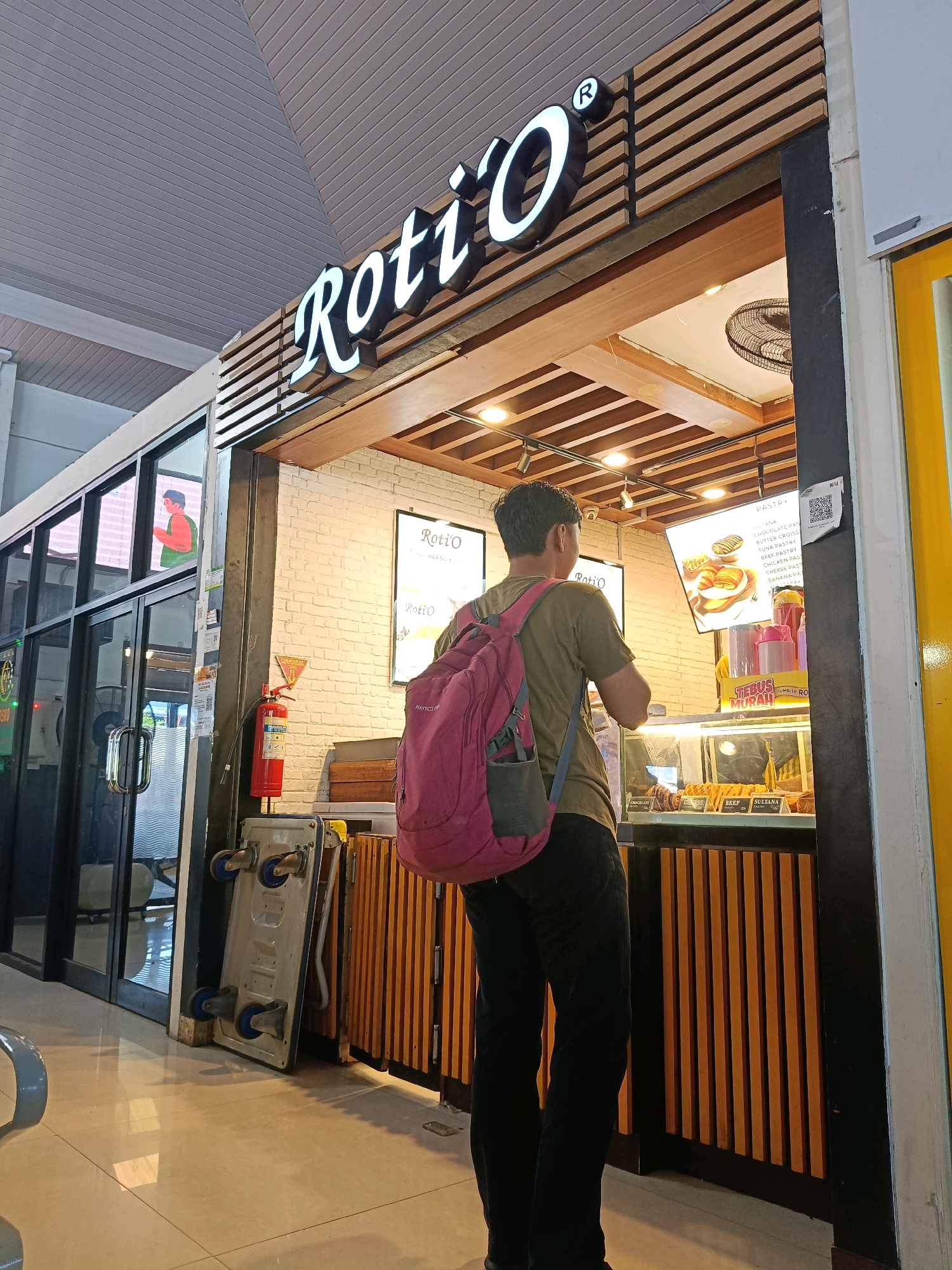 The image shows a person with a pink backpack standing in front of a Roti'O store, a popular bakery chain in Indonesia, likely located in Surabaya. The store has a distinctive wooden facade and illuminated signage. Inside the store, pasties are displayed in a glass case. A menu board above the case shows different pastry options. The floor is tiled, and the ceiling features a ribbed design. A fire extinguisher is visible on the wall to the left. The overall impression is that of a typical retail environment.