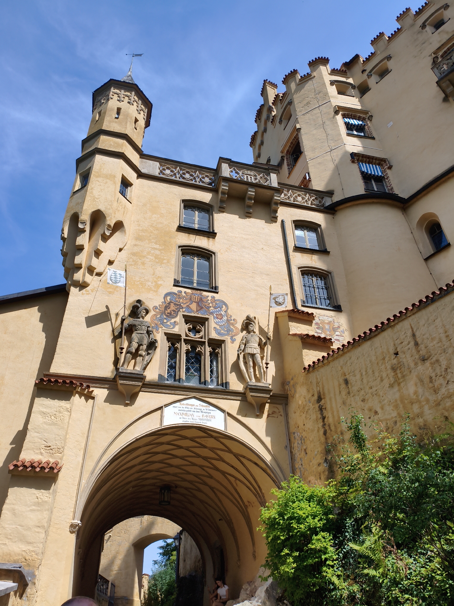 This is a low-angle, wide shot of Hohenschwangau Castle in Schwangau, Germany. The castle is a pale yellow with red tiled roofs and crenellated battlements. It is a sunny day with a clear blue sky.
In the foreground, a large arched entryway leads into the castle courtyard. Two stone statues of knights in armor stand on either side of the entryway, each holding a flag. Above the entryway is a plaque with German text. To the right of the entryway, lush green bushes and trees are growing.
The castle architecture is elaborate, with turrets, balconies, and decorative window frames. The walls appear weathered, with some cracks and imperfections. Sunlight casts distinct shadows on the castle walls, indicating its position relative to the sun.
The overall impression is one of a majestic and historic royal residence bathed in sunlight.