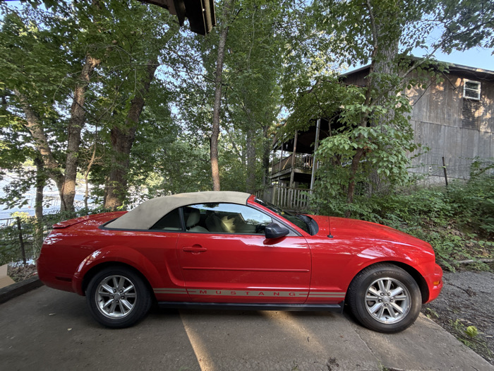 A red Ford Mustang convertible with a beige soft top is parked on a concrete surface. The vehicle features silver multi-spoke wheels and a "MUSTANG" decal with horizontal stripes on its lower side panel. The interior appears light-colored. The scene is outdoors, set within a heavily wooded environment. To the left of the vehicle, a body of water, likely a lake, is visible through a dense arrangement of mature deciduous trees. To the right and behind the vehicle, a multi-story, rustic wooden house or cabin with a wraparound porch is situated on an elevated, tree-covered slope. The sky above the tree canopy is clear blue. The location is consistent with a lakeside or forested residential area in Osage Beach, United States. There are no people present.