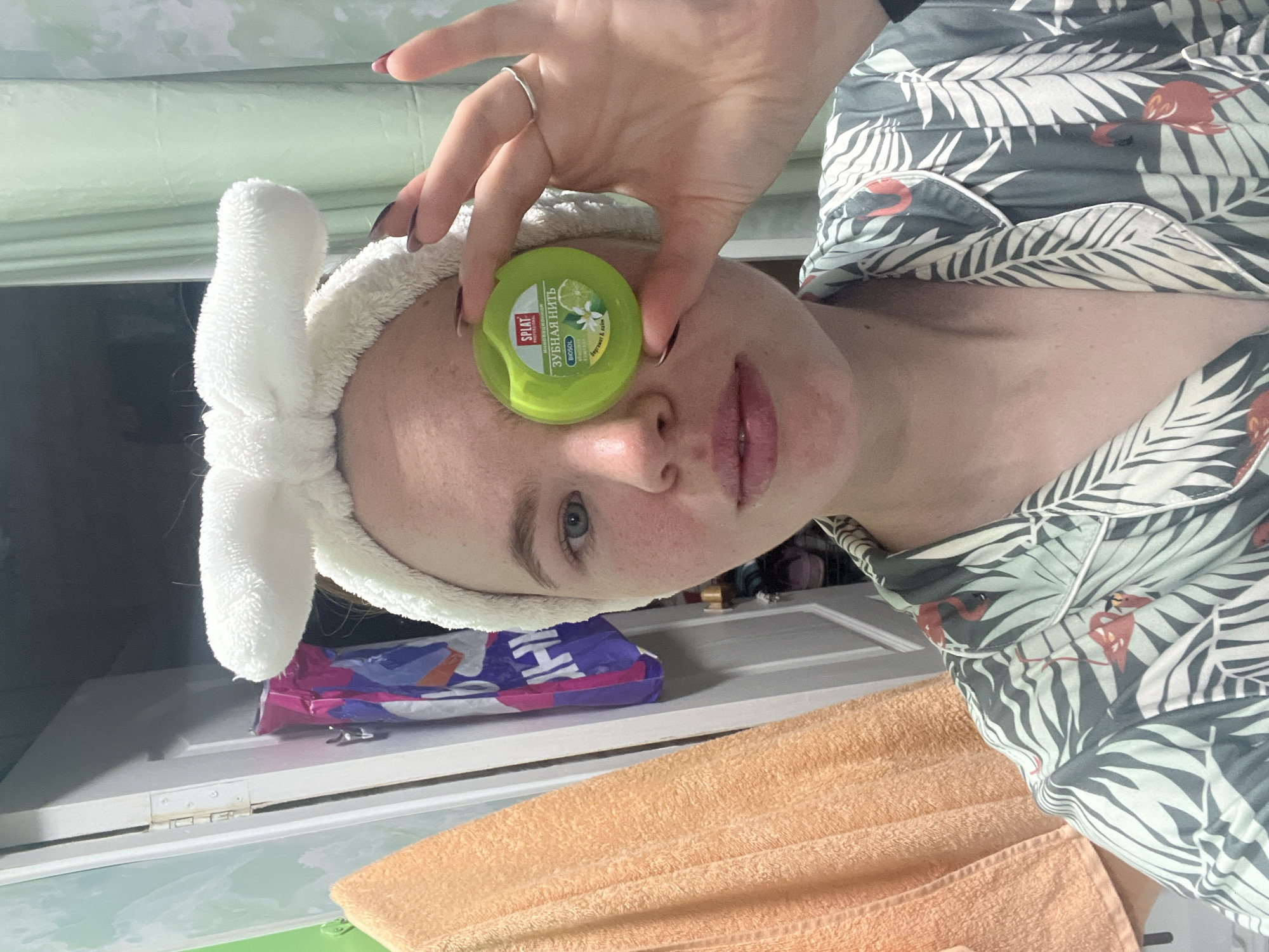 In this close-up, waist-up photograph, a young person with light skin and blue eyes is holding a small, round, green container of dental floss in front of their right eye. They are wearing a white, fuzzy headband with a large bow on top and a patterned pajama top featuring large green leaves and red flamingos. The person's left hand is visible, with dark nail polish on their fingers. The background is a slightly blurred interior scene, possibly a bathroom or bedroom. To the left, an orange towel hangs on a door or cabinet. Behind the person, there is a partially open white door, revealing a glimpse of colorful items inside, and a light green curtain. The overall lighting is soft and even. The location is identified as Chaikovsky, Russia.