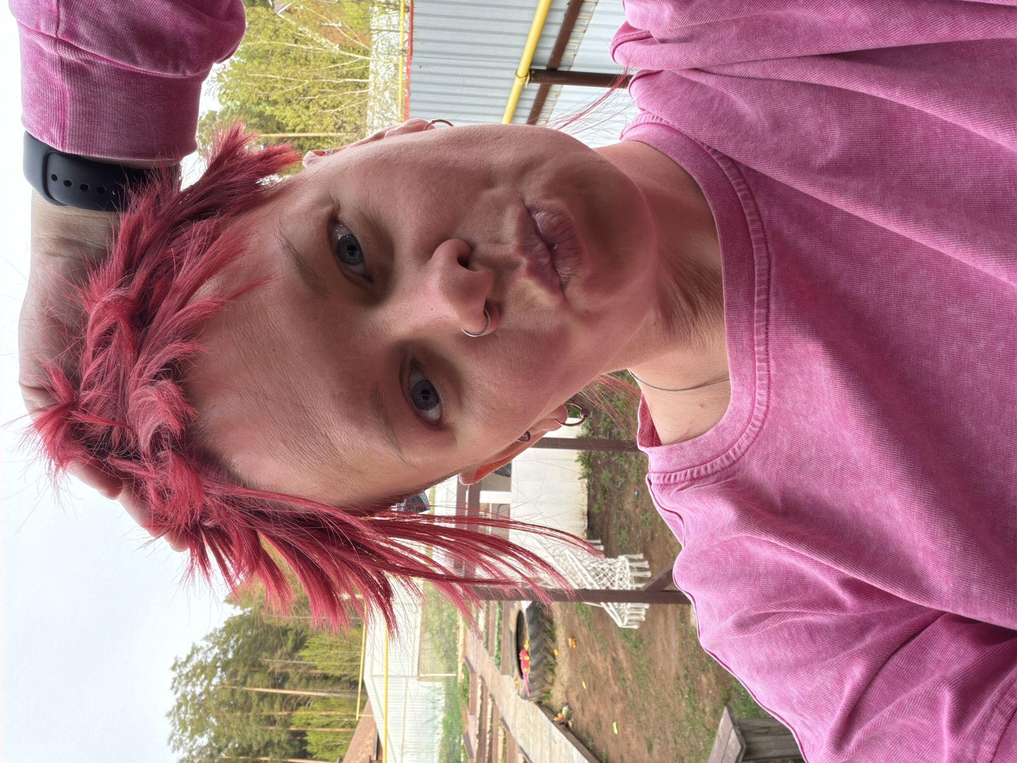 A close-up, slightly tilted, selfie-style photograph shows a person with fair skin and red, spiky hair. They are wearing a pink sweatshirt and have a silver nose ring and multiple earrings. A black smartwatch is visible on their left wrist. The person is making a kissing face towards the camera.
In the background, a rural setting is visible. There's a garden with rows of plants and a greenhouse. A tire and some wooden structures are also present. Trees are seen in the distance, and a metal roof or awning is partially visible above.
The location is Chaikovsky, Russia.