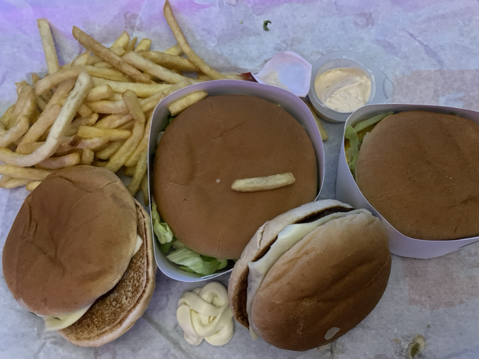 The image displays a close-up, overhead view of a fast-food meal spread out on a white, slightly crumpled paper surface, likely a tray liner or takeout wrapping. The meal primarily consists of a generous pile of french fries and multiple burgers, along with various condiments.
In the upper left portion of the image, a large mound of golden-brown french fries is visible. The fries vary in length and thickness, with some appearing crispier than others. They are scattered loosely, with some overlapping.
Towards the bottom left, a single burger is positioned, partially unwrapped or simply placed on the paper. This burger has a light brown bun, and its filling is visible on one side, showing what appears to be a breaded patty and a slice of melted white cheese.
To the right of the fries, in the center of the image, is a burger resting within a white, open-top cardboard container. The top bun of this burger is smooth and light brown. A single french fry is playfully or accidentally placed horizontally on top of this burger's bun. Below the bun, some shredded green lettuce is visible.
To the right of the central burger, another burger in a similar white cardboard container is partially visible. Only the light brown top bun and some lettuce underneath are discernible.
At the bottom center, a fourth burger is lying on its side. Its bun is light brown, and a thick, melted slice of white cheese is prominently spilling out from between the bun halves.
Several condiments are also present. In the upper right corner, there's a small, clear plastic container, partially open, holding a creamy, light-colored sauce, possibly mayonnaise or a similar dressing. Another small, white, empty plastic lid is next to it. Below this, towards the bottom center, a decorative swirl of a white, creamy sauce (likely mayonnaise or aioli) is dispensed directly onto the paper surface.
The overall impression is of a casual, indulgent fast-food meal, possibly shared among several people, indicated by the multiple burgers. The lighting suggests an indoor setting, possibly under artificial light, as there are no distinct shadows indicating natural light. No text is visible anywhere in the image.
