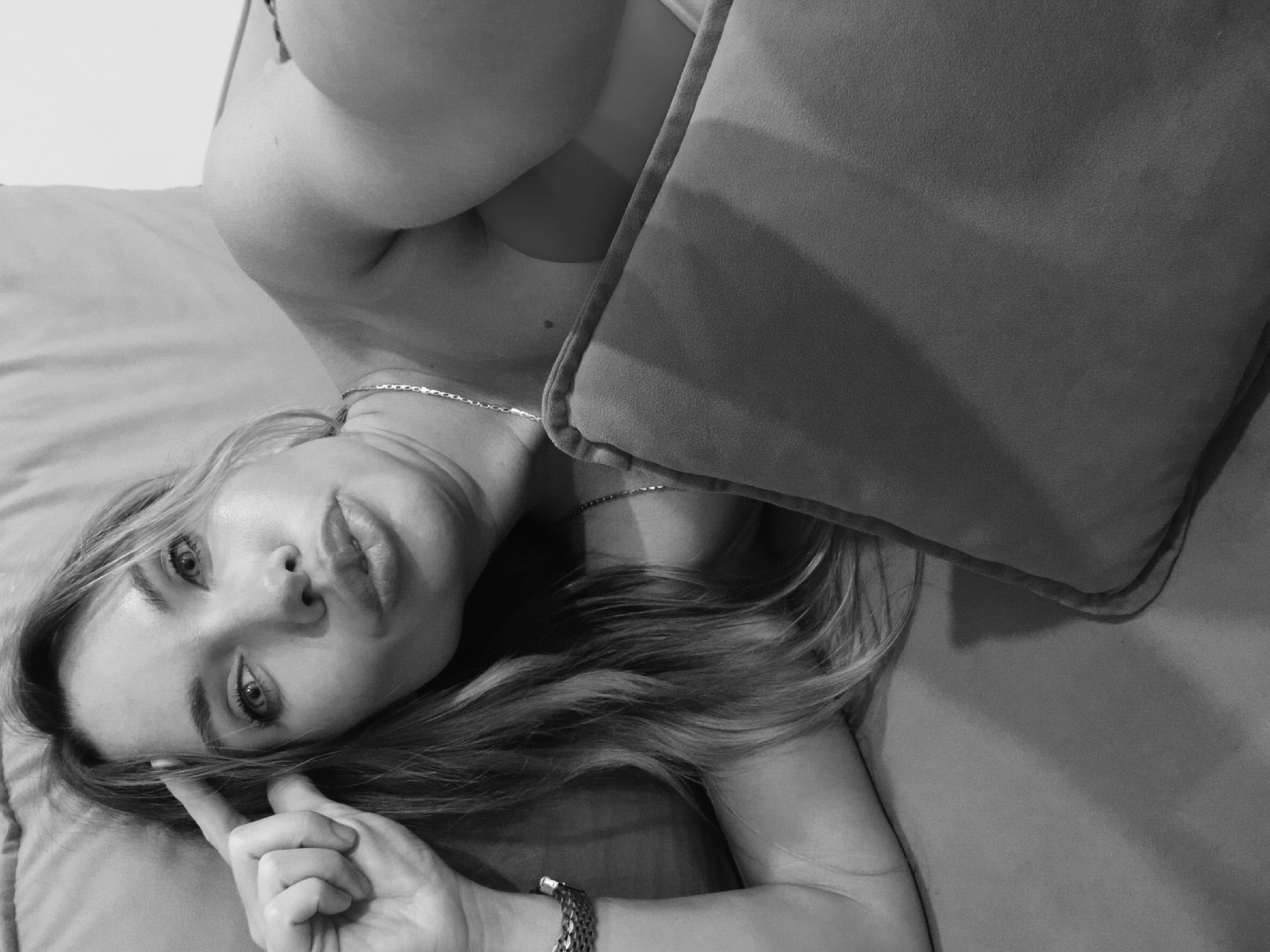 A black-and-white, eye-level, upward-facing portrait of a fair-skinned woman with shoulder-length blonde hair, lying on a bed or similar surface. She is angled towards the viewer with her head resting on the bed and a pillow beside her head on her right. She is looking up and touching her forehead with a finger on her left hand. She is wearing a bracelet, a necklace, and what appears to be a light-colored top or undergarment. The background is indistinct, showing the bedsheet or covering. The location is not identifiable.