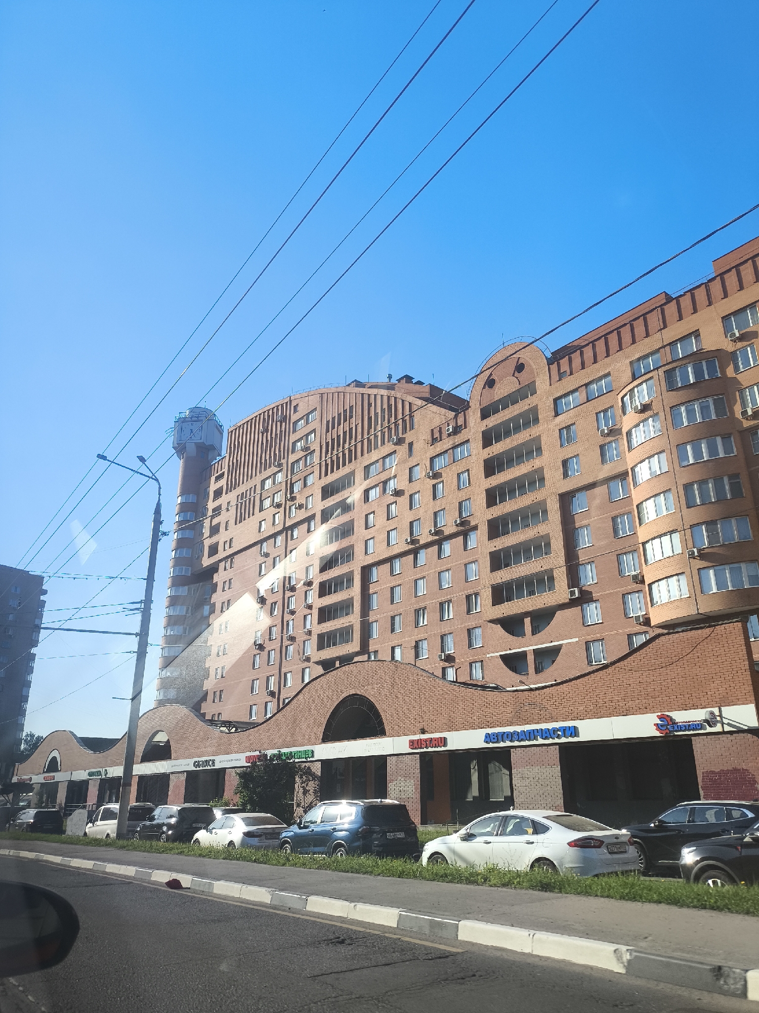 The image shows a cityscape in Unknown, Unknown on a sunny day. A tall, brick building complex dominates the view, featuring commercial units on the ground level with arched entrances. Signs in Cyrillic script are visible above the storefronts, one reading "АВТОЗАПЧАСТИ". Various cars are parked along the street in front of the building. A distinctive water tower-like structure is attached to one side of the building. Power lines run across the top of the frame against a clear blue sky. Part of a car's side mirror is visible in the bottom left corner, suggesting the photo was taken from inside a vehicle.