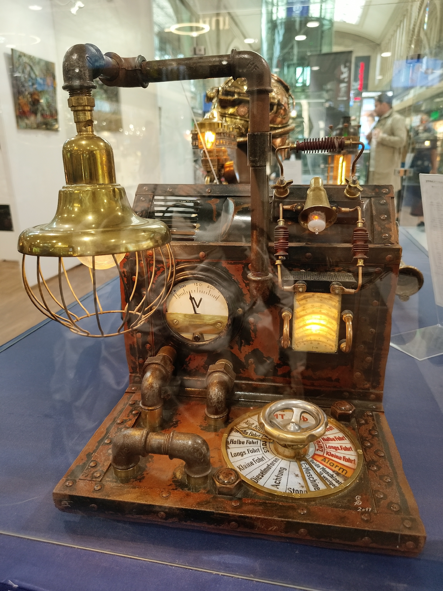 A detailed close-up shows an elaborate steampunk-style device, possibly an art piece or prop, housed within a glass display case. The device is primarily composed of rusted metal and pipes, giving it an antique, industrial aesthetic. Prominent features include a brass lamp with a wire cage, a gauge with a needle pointing towards an unknown value, and a rotary dial with settings labeled in German ("Halbe Fahrt," "Langs. Fahrt," "Kleine Fahrt," "Alarm," "Dieselmotoren," "Achtung," and "Stand"). Other elements suggest a technical or electrical function, such as what appears to be an indicator light and various connected pipes. The device is positioned on a dark blue surface within the display case. The background, visible through the glass, suggests a busy public space with people and other exhibits. There is also other art on the left of the frame. The exact location is unknown.
