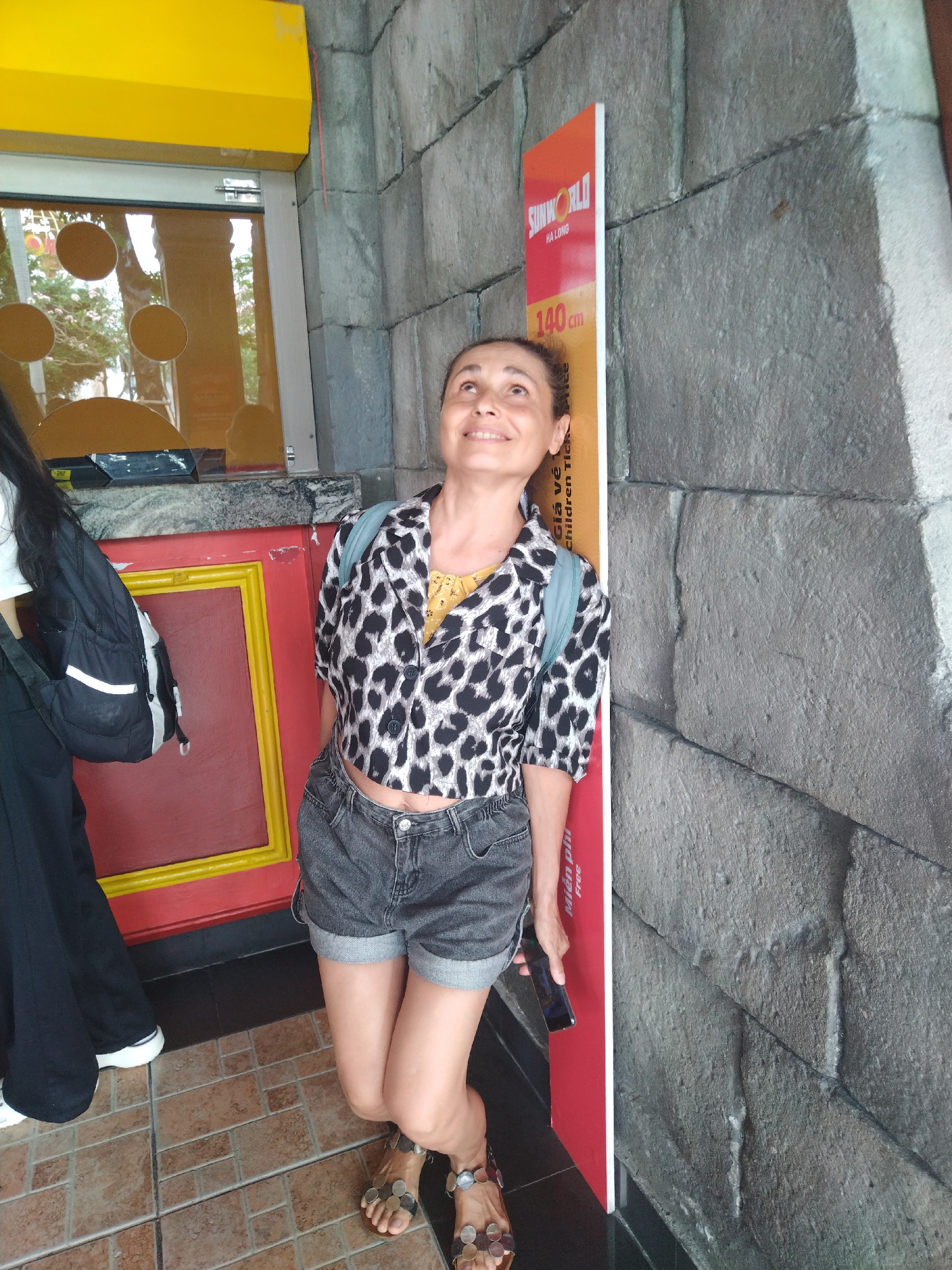 An outdoor, eye-level shot captures a woman posing next to a height measurement sign. The woman, with dark hair pulled back, is smiling broadly and looking upwards, resting against a textured stone wall. She is wearing a short-sleeved, cropped leopard print shirt over a yellow camisole, grey denim shorts, and open-toed sandals adorned with circular, coin-like decorations. A light blue backpack is visible on her shoulders, and she holds a dark smartphone in her right hand.
To her right, a red and white vertical sign features the logo "SUNWORLD HA LONG" and the measurement "140 cm." Partially visible text below includes "Giá vé" and "Children Tick Free," along with "Miễn phí." This sign appears to be a height guide, likely for admission purposes at an amusement park or attraction.
To her left, partially obscured by another person's dark clothing and black backpack, is a ticket window or counter. This area has a bright yellow awning above a window that reflects trees and circular patterns. Below the window, there's a red structure framed with yellow trim. The background wall behind the woman and the sign is made of large, irregularly shaped grey stone blocks. The ground is a tiled floor with light and dark brown squares. The bright lighting suggests it is daytime. The woman's expression conveys happiness and amusement.
