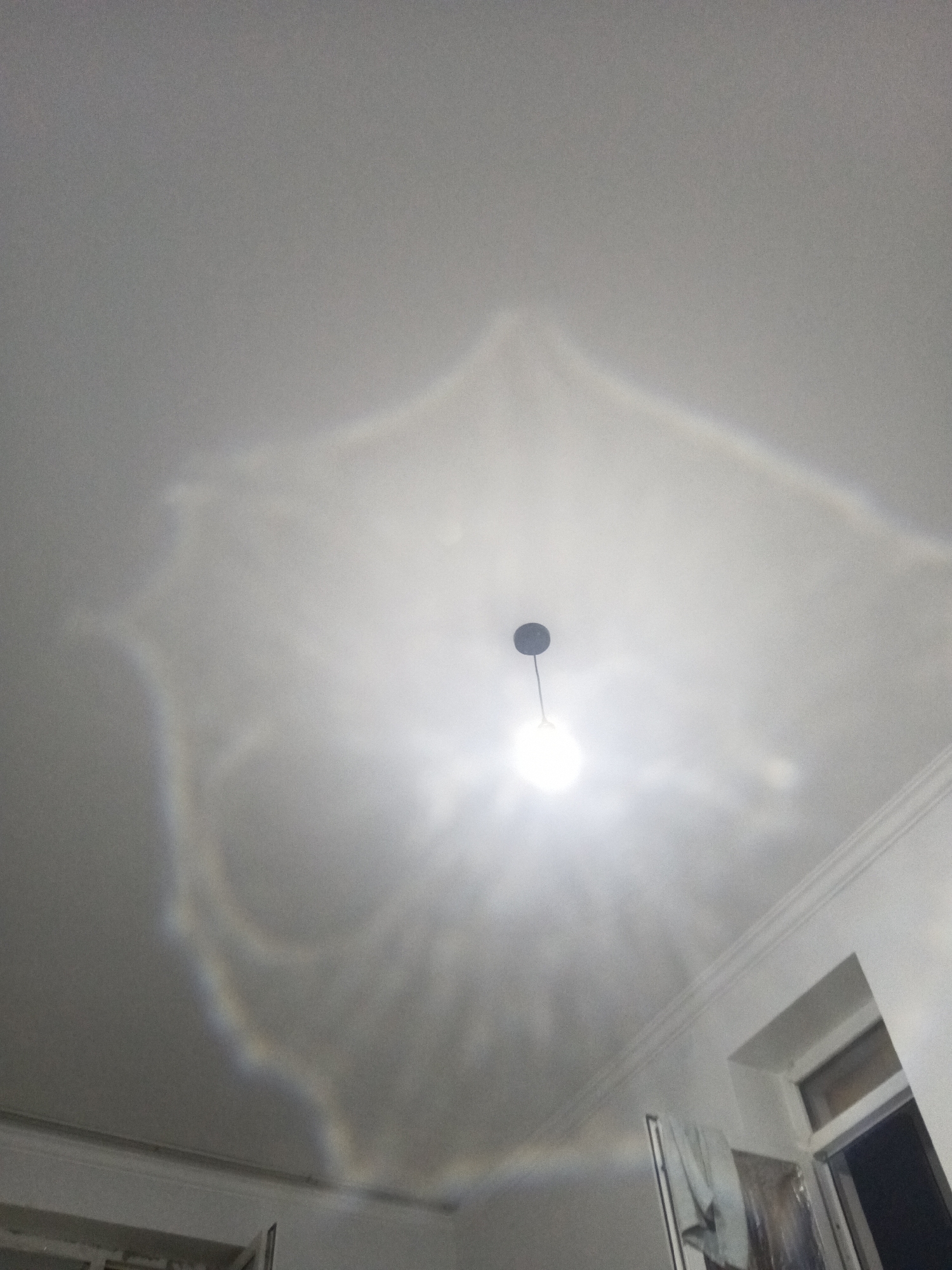 The image shows a white ceiling in a room, with a light fixture hanging from the center. The light fixture has a round, bright bulb. Around the light fixture, there are iridescent patterns on the ceiling, possibly caused by light refraction. The ceiling has crown molding along the edges. In the lower right of the image, a portion of a wall with a window frame and molding is visible. The overall impression is of an indoor scene, possibly an apartment or office, illuminated by the central light fixture, with unusual light effects on the ceiling. There are no people or specific identifiable landmarks that would indicate the city of Astana, Kazakhstan.