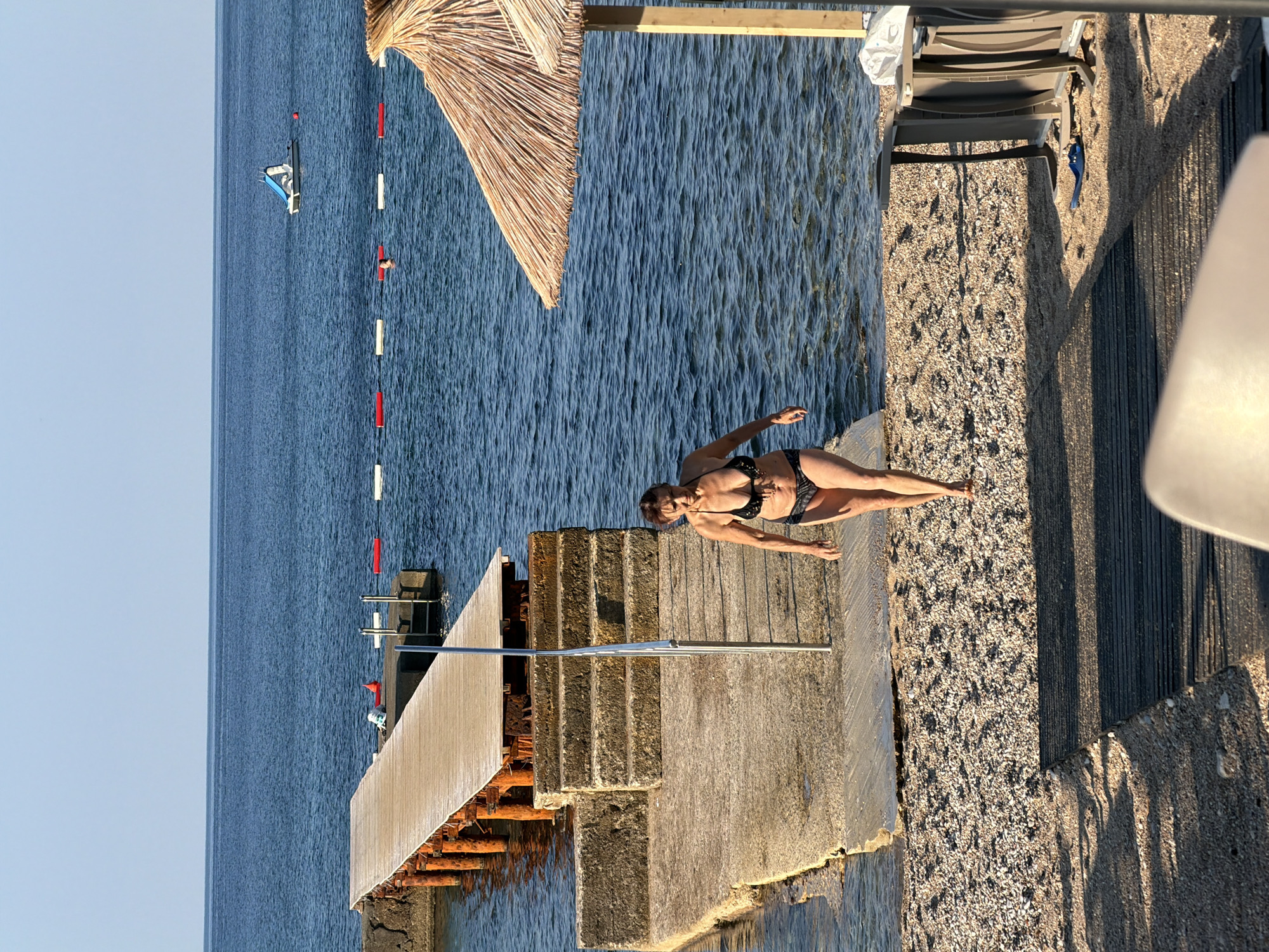 A woman in a black bikini walks on a pebbled beach towards the foreground of the image. She is emerging from the water and walking on a concrete path that leads away from a wooden pier. The pier extends into the calm, blue sea, supported by orange metal pilings. In the background, the sea stretches to the horizon under a clear, light blue sky. Buoys mark a swimming area in the water, and a small white pedal boat with a blue slide is visible in the distance. A straw umbrella provides shade on the beach to the right of the image, with a lounge chair and other beach furniture nearby. The scene appears to be a coastal resort area, possibly in Budva, Montenegro, given the general aesthetic of the beach, pier, and surrounding water.