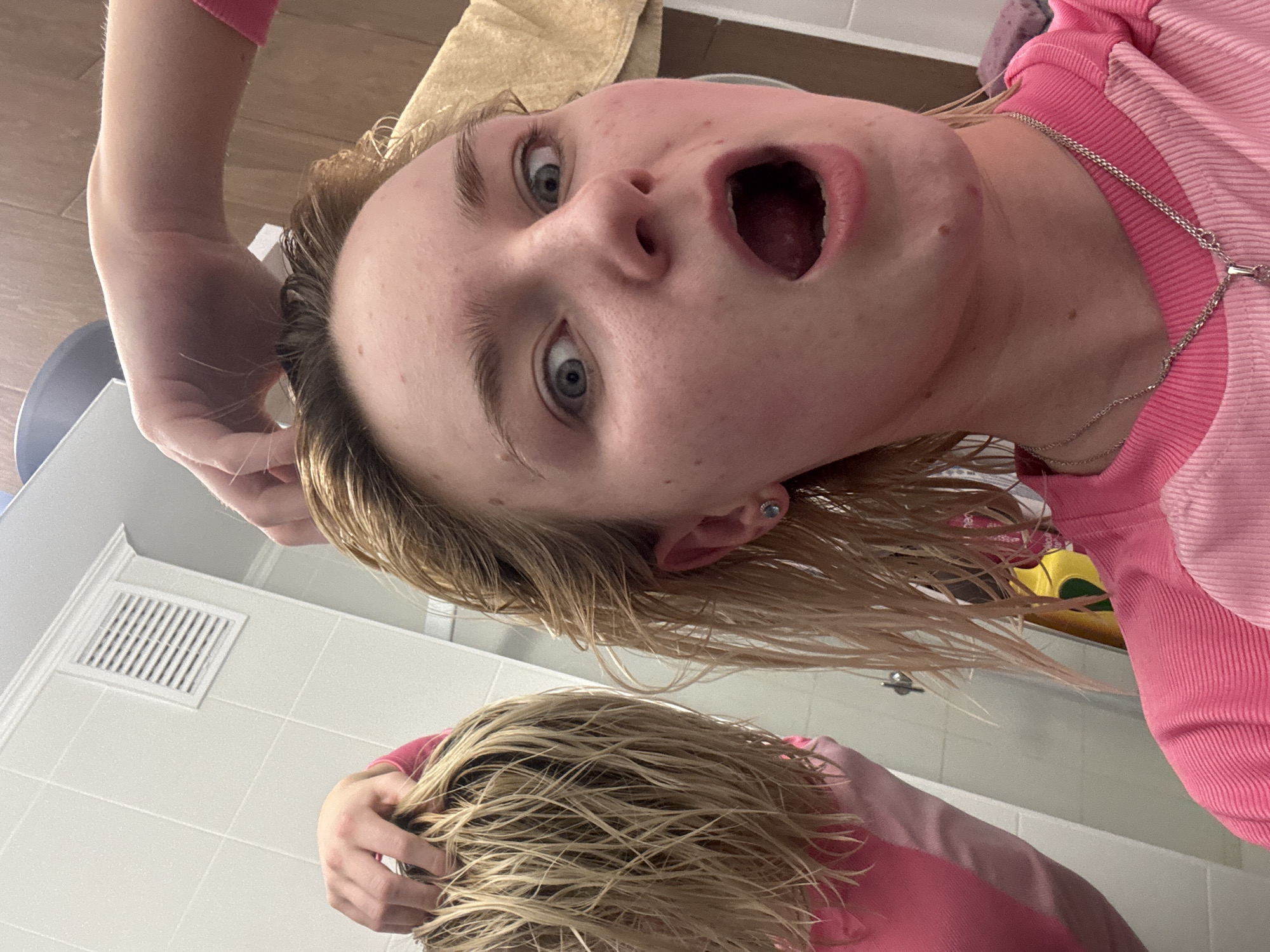 A young individual with light blonde, wet hair and wide blue eyes is the central subject of this close-up, rotated image. They are wearing a pink ribbed long-sleeved shirt and a silver chain necklace, with a small silver earring visible on their left ear. Their mouth is open wide, conveying an expression of surprise, awe, or playful shock.
The person appears to be looking into a mirror, evidenced by a distinct reflection of their wet hair and upper torso, including part of their pink shirt, in the lower section of the frame. One of their hands is visible touching their wet hair on the left side of their head, and a reflected hand is also seen in the lower part of the image, similarly touching hair.
The setting is an indoor space, likely a bathroom, indicated by white tiled walls with visible grout lines and a white rectangular air vent/grille mounted on the wall. A beige towel is draped in the upper right background. Partially visible objects in the reflection include yellow and green items, and a metal fixture that could be part of a sink or shower. The unusual, low-angle perspective, combined with the image's rotation, creates a dynamic and somewhat disorienting view, suggesting the photo was taken while looking upwards, possibly from a sink or bath. The wet hair implies recent washing. No visible text is present in the image. The location context is Ligolambi, Russia.