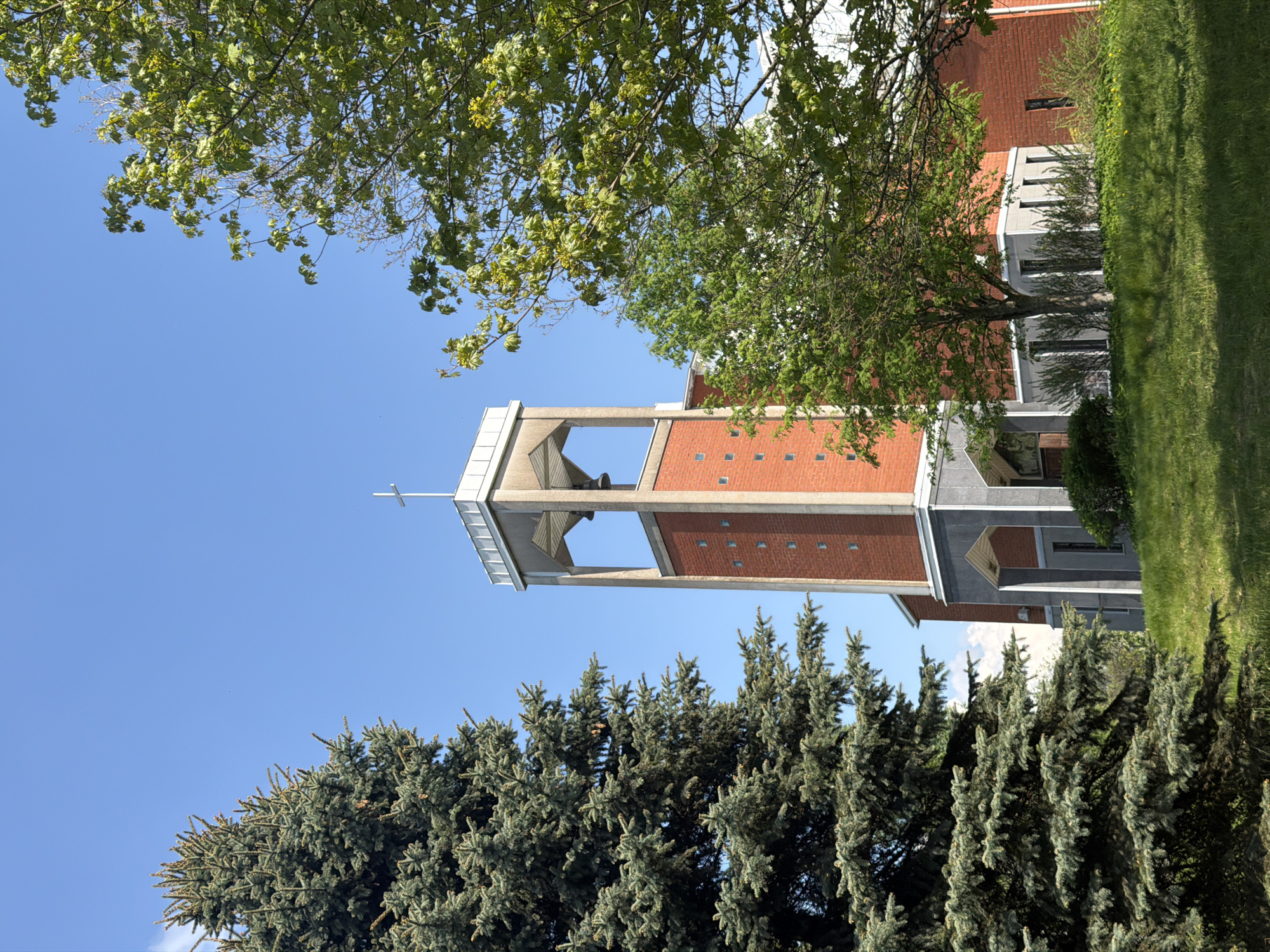 The image depicts a building, possibly a church, with a tall, rectangular bell tower featuring a cross at the top. The tower is constructed of red brick with concrete or stone accents. The building is framed by trees with green foliage at the top and blue spruce trees at the bottom. The sky is clear blue. The scene suggests a sunny day. The location in the city of Tokkari, Russia, is not apparent from the visual information provided.
