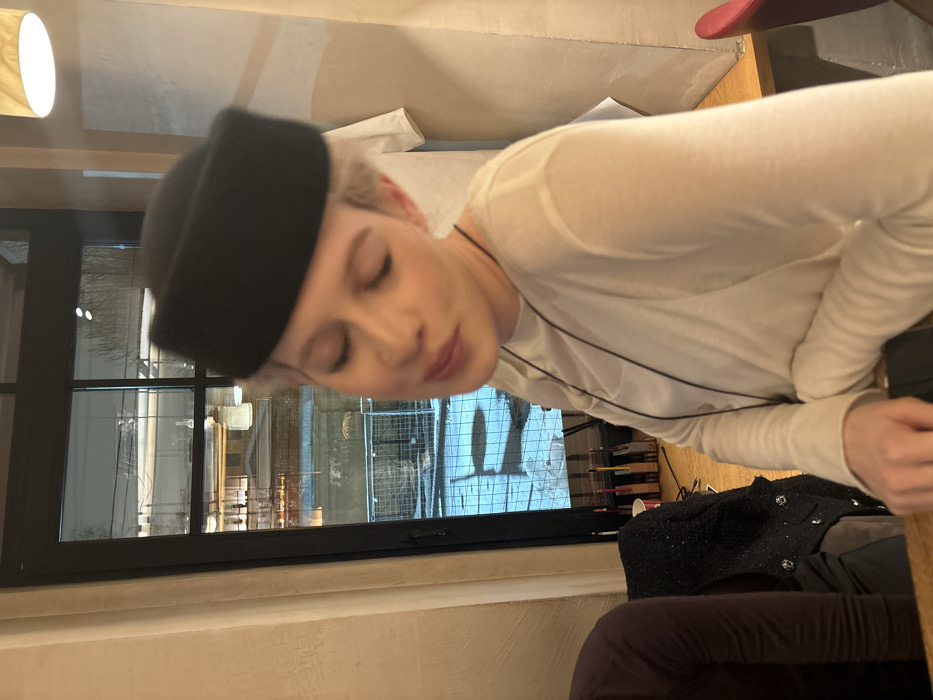 A woman wearing a black hat and a white long-sleeved shirt looks down with her eyes closed. A long black string hangs around her neck. She is sitting at a table, with a blurred view outside a window. The scene includes books, a dark-colored jacket, and a purple garment. The overall image is slightly tilted.