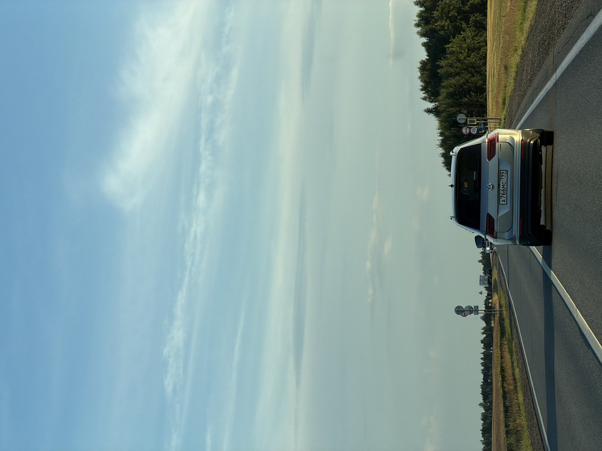 An eye-level, medium shot captures the rear of a silver Volkswagen Tiguan driving on a paved road, heading away from the camera. The car's license plate reads "A766HC 799 RUS". The road is a two-lane highway with a white dashed line dividing the lanes and solid white lines marking the edges. The highway curves slightly to the left. On the right side of the road, a grassy shoulder is visible, followed by a field of dry, yellowish grass. Beyond the field, a dense forest of green trees forms the horizon. To the left of the road, another field of dry grass stretches out, and in the distance, a few cars and a truck are visible on the highway. There are several road signs on the left side of the road, including a sign with three red circles and a sign indicating a speed limit of 70. The sky above is a pale blue, with scattered wispy white clouds. The scene appears to be a rural setting, possibly on the outskirts of the city of Obukhovo, Russia, given the license plate. The lighting suggests it is late afternoon or early evening.