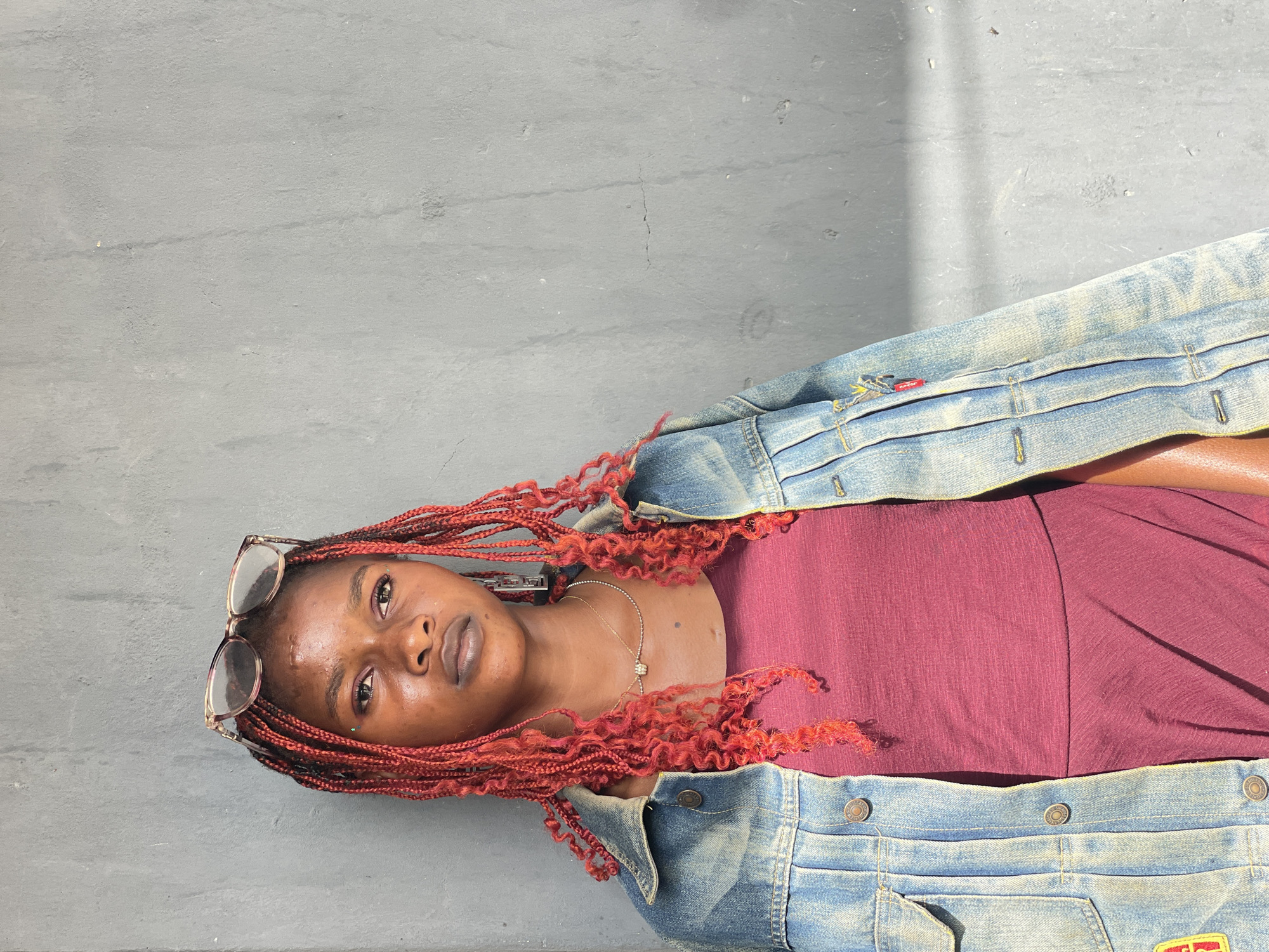 The image features a young woman with dark skin and vibrant, reddish-orange braided hair, lying horizontally on a textured grey surface, possibly concrete or asphalt. She looks directly at the viewer with a composed, neutral expression. Her long braids, some with curly ends, are spread around her head and shoulders.
She is dressed in a casual outfit, consisting of a deep burgundy-colored top or dress, and an open, light blue denim jacket. A delicate gold-colored necklace with a small, sparkling pendant is visible around her neck. A pair of tortoiseshell-framed sunglasses is perched on top of her head, resting on her braided hair. On the left chest pocket of her denim jacket, a small red tag with white lettering, appearing to be the "LEVI'S" brand logo, is visible.
The background is a uniform, plain grey, suggesting an outdoor location under bright daylight. Subtle shadows on the surface indicate the sun's position. The scene depicts a posed individual in a casual setting in Badagry, Nigeria.