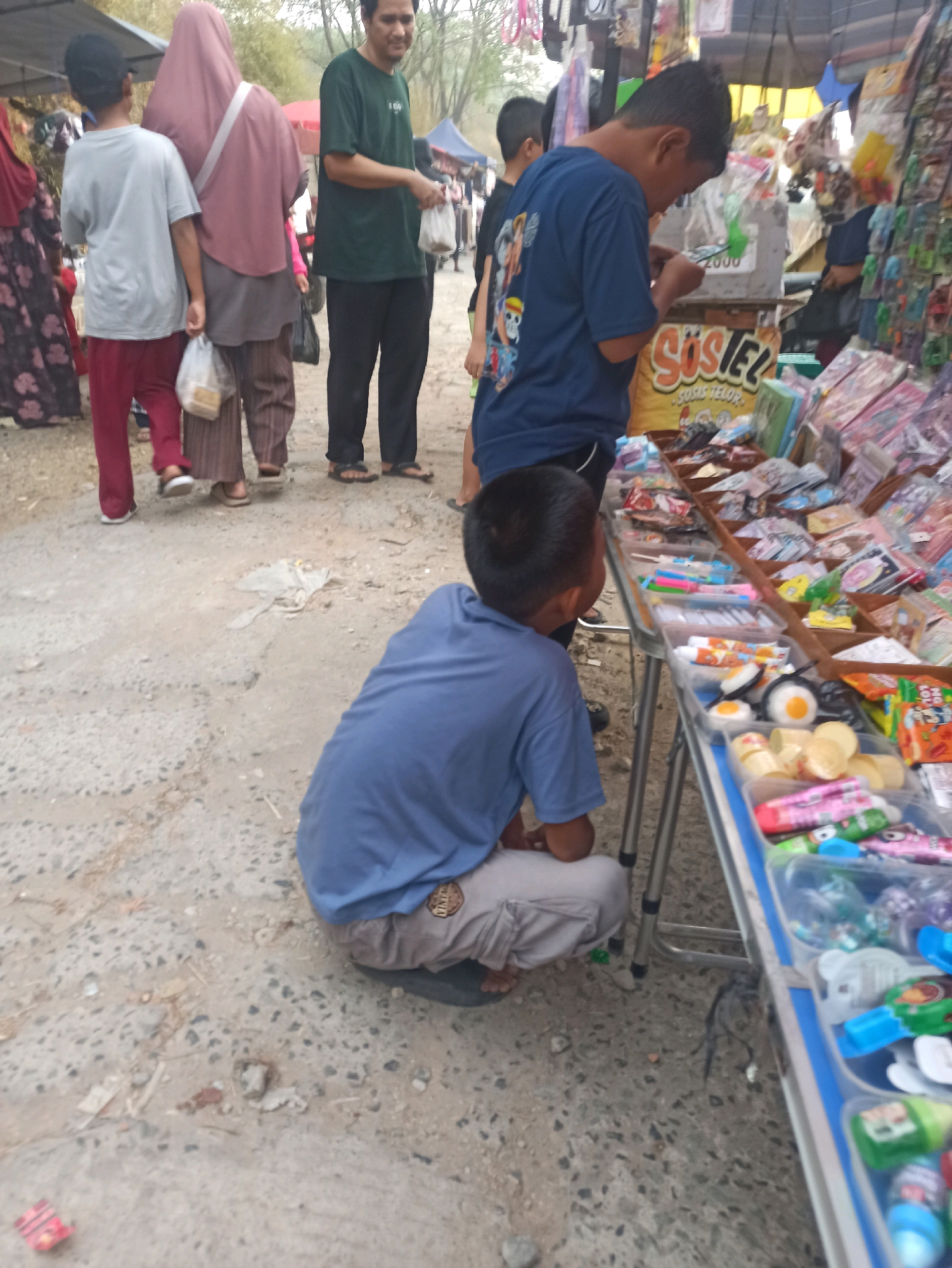In Telagamurni, Indonesia, a busy outdoor market is depicted. Several individuals are present, including a boy in a blue t-shirt and light-colored pants crouching down, examining items on a table laden with various snacks and toys. Next to him, another boy in a blue shirt stands looking at the table. Further back, a man in a green t-shirt and black pants, holding a plastic bag, converses with a woman wearing a pink headscarf and a darker colored outfit, who is also carrying a plastic bag. Another person in a red shirt and red pants is visible to the left. The ground is a mixture of dirt and gravel. The market stalls display a colorful assortment of packaged goods, including what appears to be candy and small toys. The overall scene suggests a typical market day with shoppers and vendors.