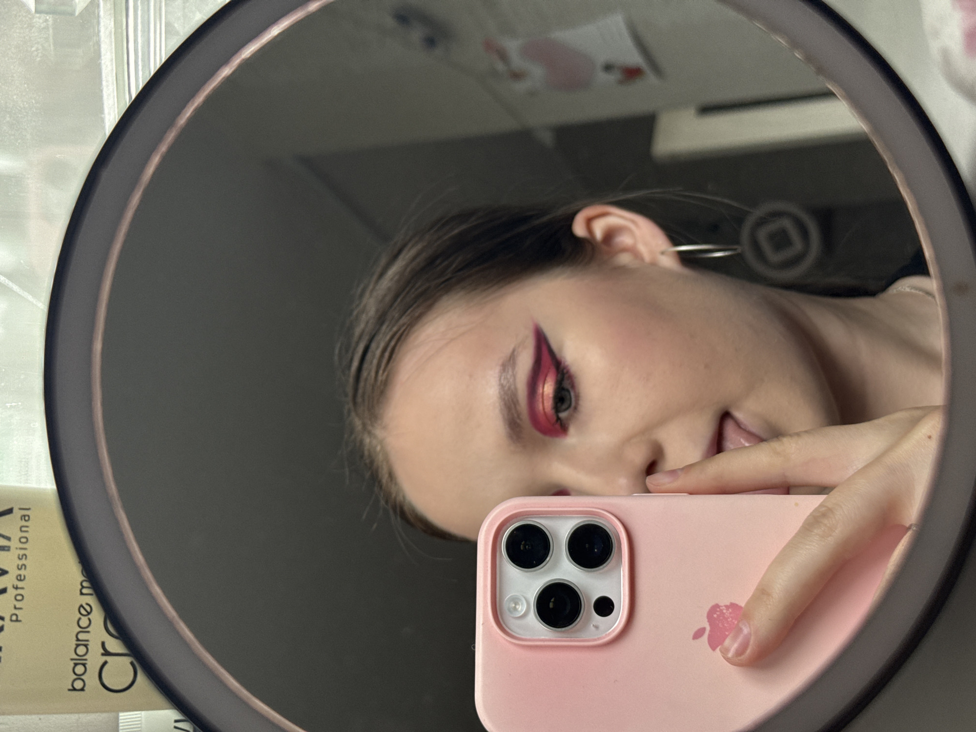 A young woman is taking a selfie in a round mirror with an iPhone. The phone has a light pink case with a faded floral design near the bottom. The woman has dark hair and wears red and pink eyeshadow. Her right ear is visible and has a hoop earring. The reflection in the mirror shows a glimpse of the room behind her, including a white surface and a sticker. In the foreground, to the left of the mirror, there's a tube of "Kravia Professional balance" cream. There's no clear evidence that this photo was taken in Chaikovsky, Russia.