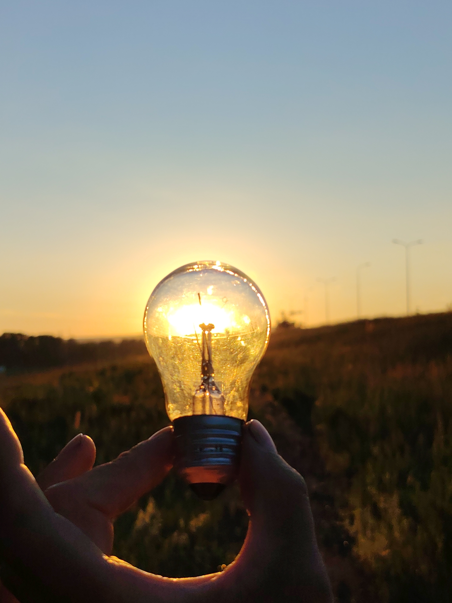 A hand holds an incandescent lightbulb up to a sunset sky. The lightbulb is positioned in the foreground and is translucent, allowing the golden light of the setting sun to shine through it. The sun is visible behind the lightbulb, creating a bright, warm glow. The background consists of a rural landscape with trees and fields. In the far distance, there are several tall streetlights, suggesting the presence of a road or urban area. The sky transitions from a pale blue at the top to a warm orange and yellow near the horizon. The image was taken during the golden hour, with the low angle of the sun casting a soft light. The location is identified as Chaikovsky, Russia.