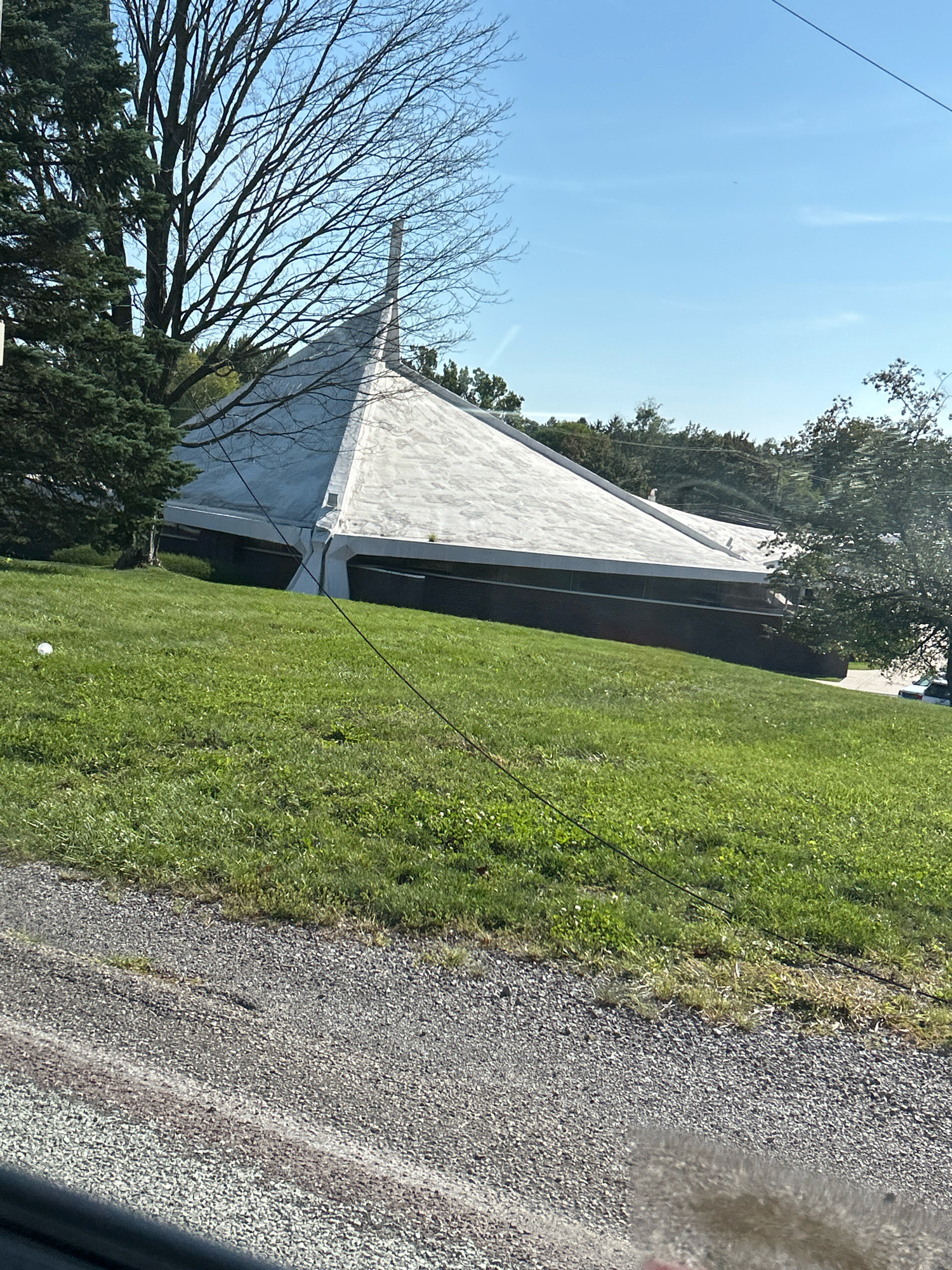 The image shows a large, angular building with a white, peaked roof, situated on a grassy slope. The building has dark, horizontal windows. A tall, bare tree stands to the left of the building, and evergreen trees are also visible. A dark wire runs diagonally across the foreground and over the roof of the building. The ground in the foreground is a gravel surface. The scene appears to be outdoors on a sunny day. The building's unique architecture suggests it could be a place of worship or a public building. The context of Robinson Township, USA, indicates a suburban or semi-rural setting.