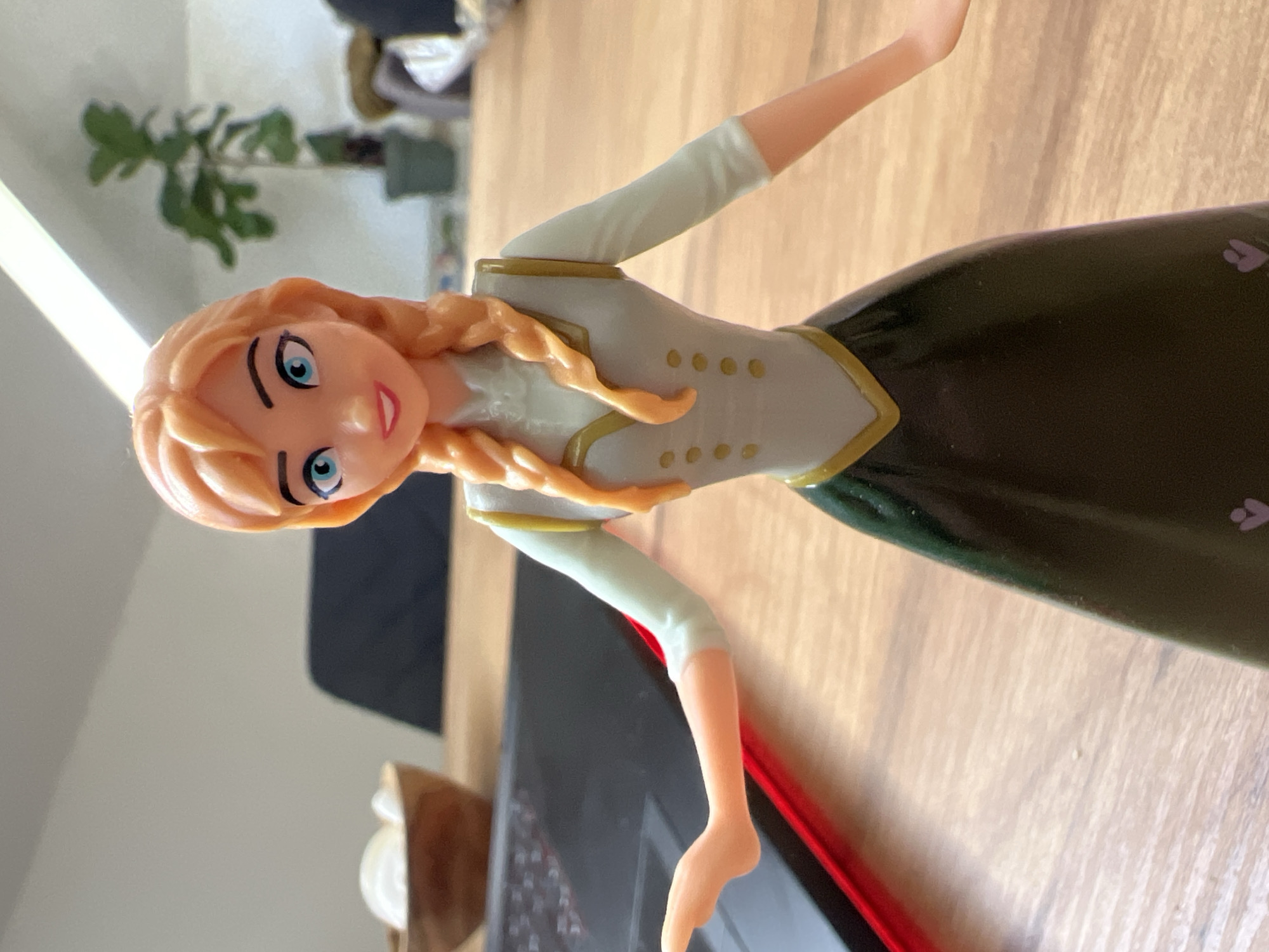An Anna figurine from Disney's Frozen franchise is shown in a medium close-up shot. The figurine has orange braided hair, blue eyes, and is wearing a light blue long-sleeved shirt with a grey vest adorned with gold trim and four gold buttons. Her skirt is dark green. She is standing on a wooden table. In the background, a dark laptop computer with a red edge is visible, as well as a wooden bowl containing white objects, a potted plant, and a wall. The image is taken indoors, likely in a home or office setting in Saint-Vith, Belgium, given the context.