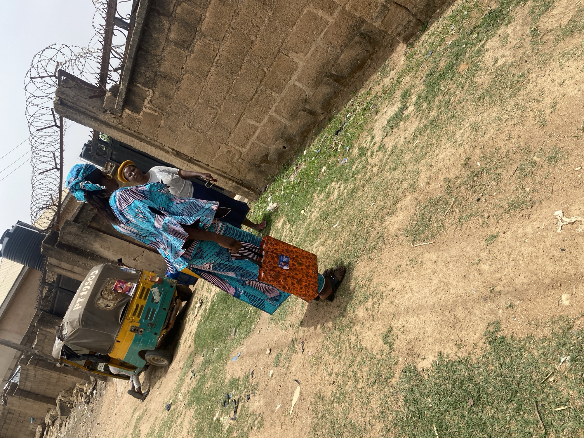 This image captures an outdoor scene in Barakin Akawo, Nigeria, under clear daylight conditions. Two women are prominent subjects on a dirt path mixed with sparse green grass.
In the foreground, a woman is seen from a side-rear perspective, dressed in a vibrant blue and patterned traditional outfit, complete with a matching headwrap. She is holding a rectangular, orange-brown package adorned with a visible but unreadable pattern or logo. Her footwear consists of brown sandals.
Behind her, another woman stands, wearing a white t-shirt, dark trousers, and a yellow head covering. She is looking towards the viewer with a smile, holding what appears to be a small item in her right hand.
The background features a sturdy, light brown wall, possibly constructed from stone or concrete blocks, which is topped with security razor wire. To the left, a yellow and green three-wheeled auto-rickshaw, commonly known as a Keke Napep, is visible. Its license plate, "PT-19503," is clearly discernible. Further behind, part of a building and a black water tank on a stand are faintly visible. The ground is unpaved, indicating a street or open area. The clear sky suggests good weather.