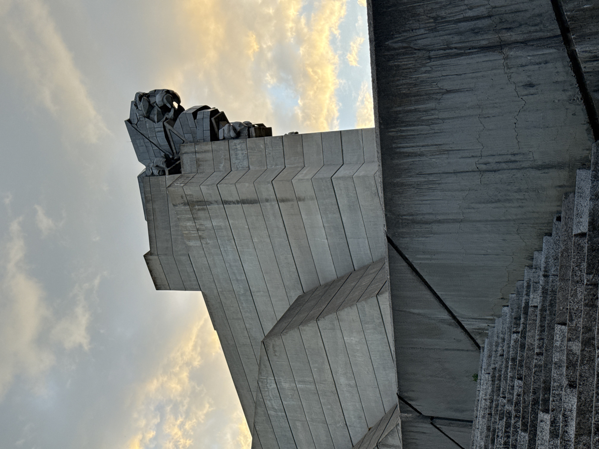 The image depicts the Monument to 1300 Years of Bulgaria, located in Shumen, Bulgaria. It's a large, abstract concrete structure consisting of geometric shapes and a sculptural group at the top. The sky is visible in the background. The image is taken from below, looking up at the monument. Steps are also visible on the right.