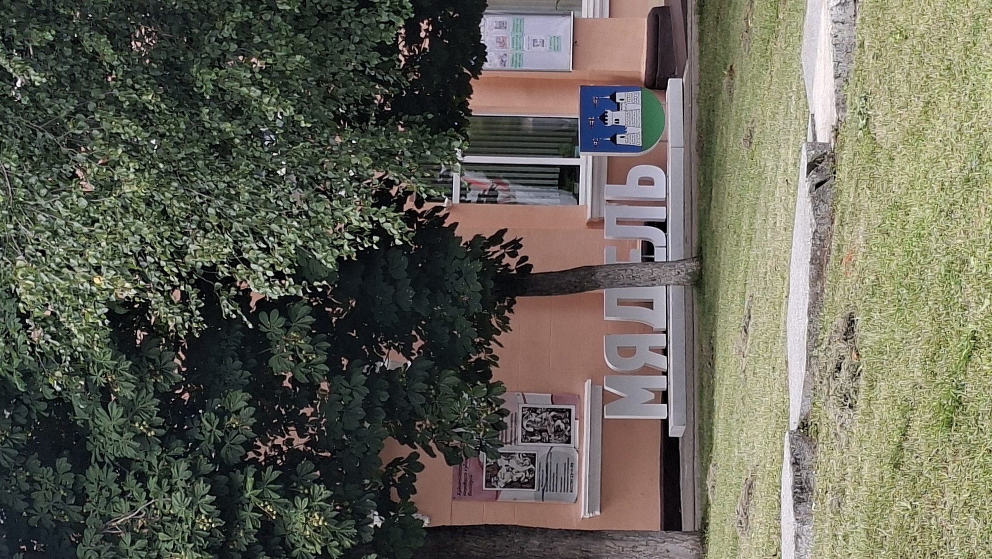 The image shows a street-level view of a building with a peach-colored facade. In front of the building, large white three-dimensional letters spell out "МЯДЗЕЛЬ" (Myadzyel). To the right of the letters, a coat of arms featuring a castle on a green hill is displayed. The building has several windows, some with blinds. A tree with green leaves partially obscures the facade and the letters. To the left of the letters, a framed poster with images and text is mounted on the wall. In the foreground, a grassy area with some paved elements is visible. The overall setting appears to be an outdoor public space.
