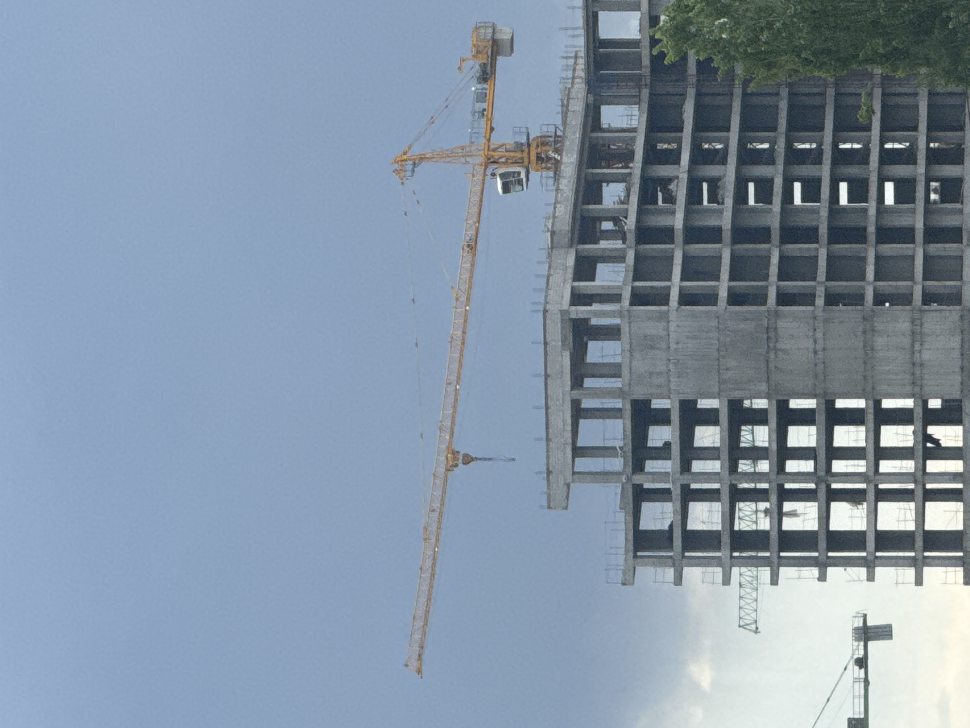 An exterior shot features a concrete structure under construction alongside two construction cranes. The structure has a grid-like facade with open spaces for windows. A tall, yellow construction crane stands beside the structure. In the lower right, there is a second construction crane partially visible. The sky is a light blue, with some cloud cover near the bottom right. Green trees are visible at the top right corner. This image was taken in Yerevan, Armenia.
