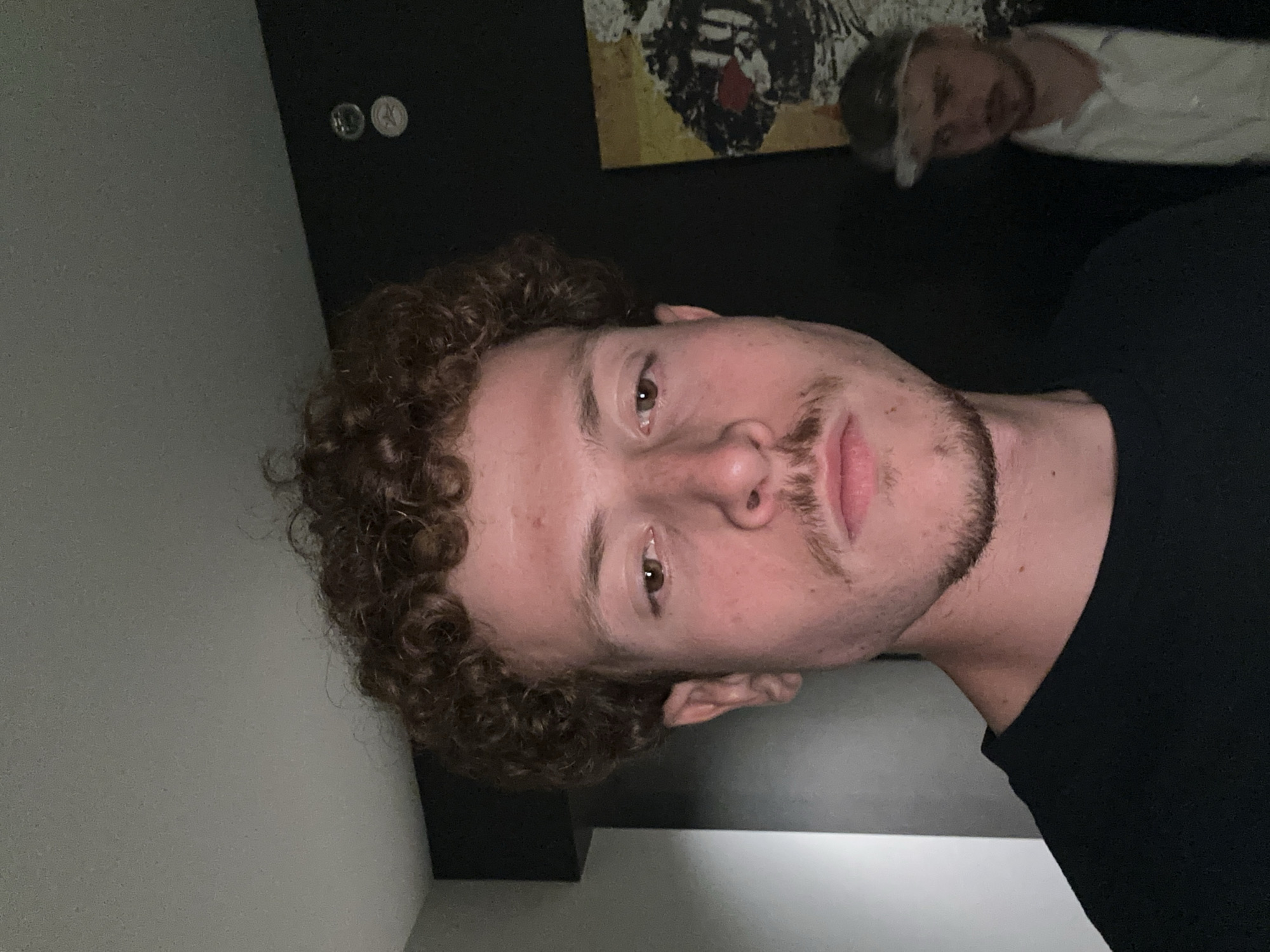 A man with curly brown hair and a mustache is facing the camera. He is wearing a black shirt. Behind him is a dark wall with two round objects mounted near the top. A piece of artwork is also visible behind him. Another person is partially visible in the background. The location is not identifiable as College Park, United States of America from the available visual information.