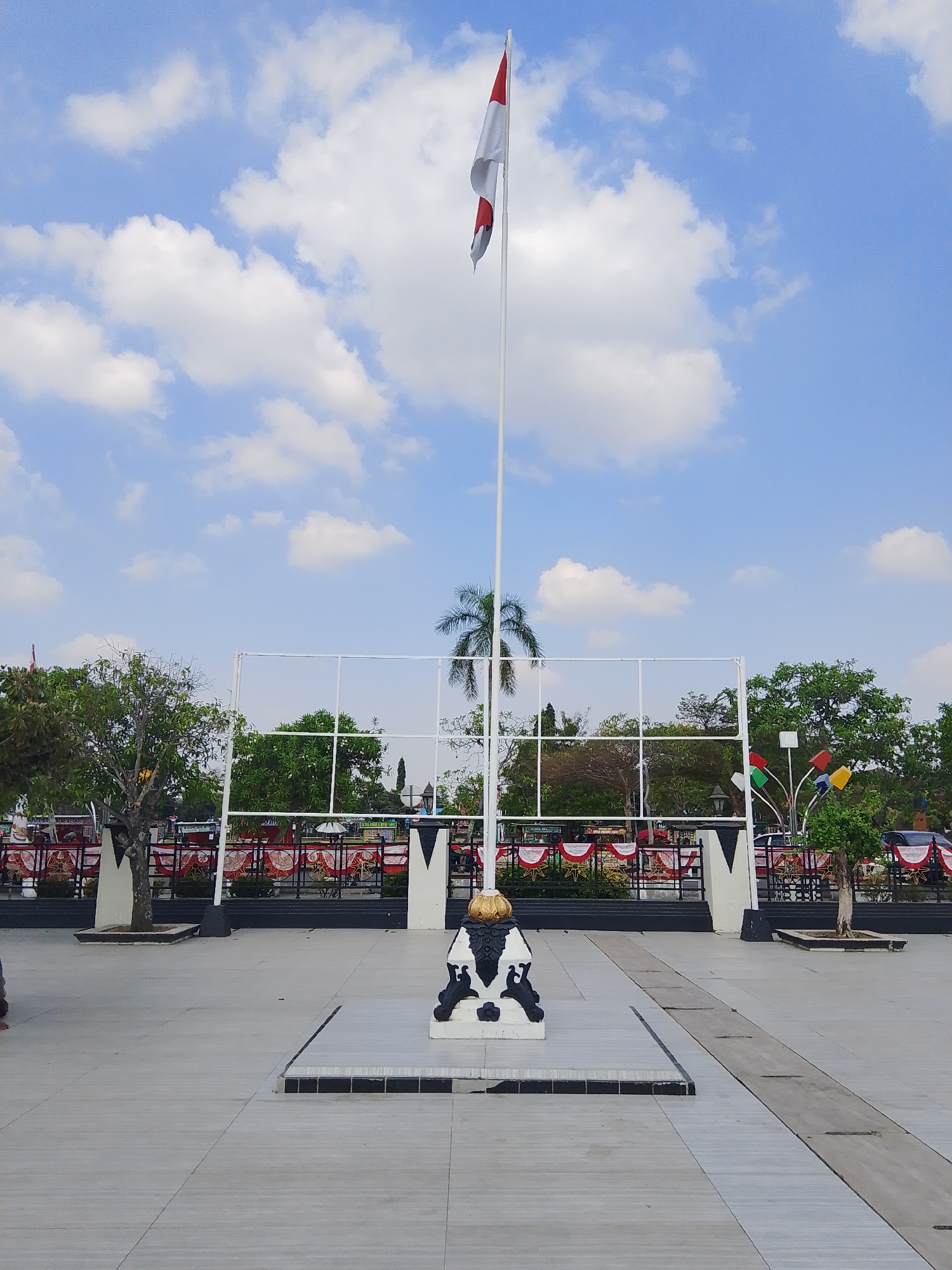 The image depicts a wide, open paved area in front of a park or public space. A tall flagpole stands prominently in the center, flying a red and white flag, likely the Indonesian flag. At the base of the flagpole is a decorative sculpture featuring a golden orb on top of a black and white statue.
Behind the flagpole, a white metal fence or structure with rectangular openings forms a boundary. Beyond this, a lush green park is visible with various trees, including a palm tree. Red and white festive banners are strung along the fence. In the distance, more trees and some structures can be seen. The sky is a bright blue with scattered white clouds.
The paved area itself is laid with light-colored square tiles. A dark strip runs diagonally across the pavement in the foreground.
The presence of the Indonesian flag and the likely setting of a public park suggest this location is within Indonesia. Given the description of the scene, this could be a town square, a monument site, or an area commemorating national holidays in a city like Demak, Indonesia, known for its historical significance.
There are no people clearly visible in the image. The overall scene is tranquil and patriotic, set under a clear sky.