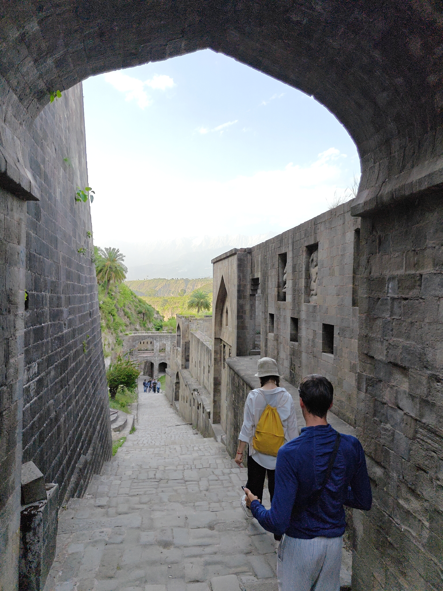 Two people walk down a stone-paved street bordered by high stone walls. The wall on the right features recessed windows with sculpted figures. The street descends through an archway that frames a view of green hills. In the distance, a group of people are walking in the same direction down the street. One person has a yellow backpack. The location is in Kaswara, India.
