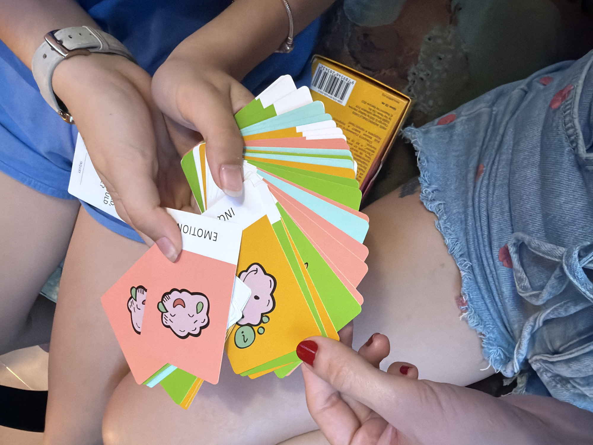 The image shows a close-up view of two people holding a fan of colorful cards. The cards have cartoonish illustrations on them. One card is pink and shows two small, pink blobs with green accents and what appear to be hands. Another card is yellow and depicts a similar pink blob with a question mark above it. The cards are fanned out, revealing a variety of colors including pink, green, and light blue on their edges. A yellow box with a barcode and some text in a foreign language is visible at the bottom right of the image. One person's hand, with red nail polish, is holding the cards. Another hand, wearing a gray watch, is also visible. The individuals are wearing casual clothing, including denim shorts with red polka dots. The background is out of focus but appears to be an indoor setting. There is no visible indication of the location being Svilengrad, Bulgaria.