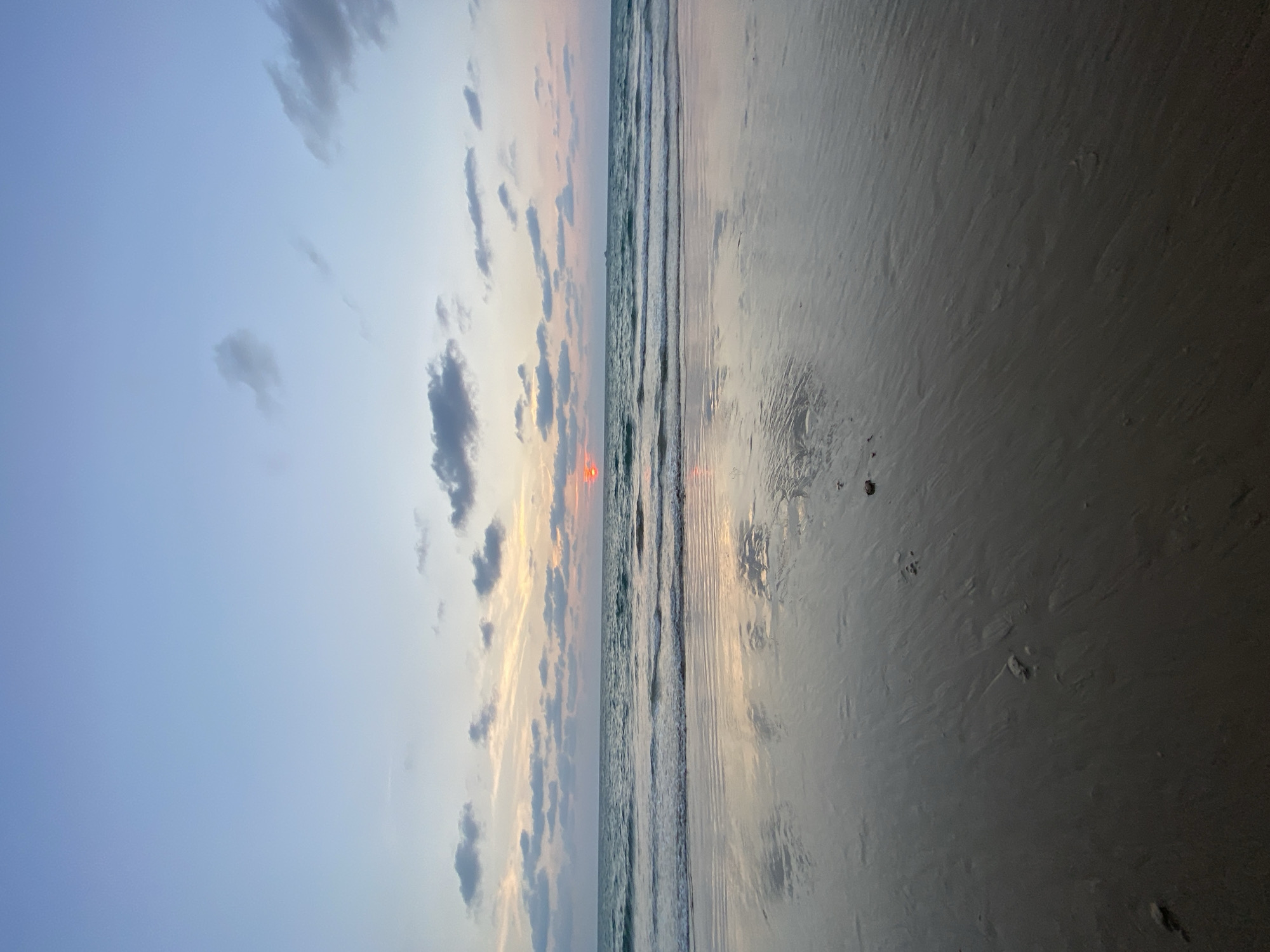The image depicts a beach scene at sunset in Choeng Thale, Thailand. The sky exhibits a gradient of colors, transitioning from a clear blue to a pastel mix of yellows, oranges, and pinks near the horizon. Clouds are scattered across the sky, some dark and dense. The sun is visible near the horizon, appearing as a bright orange orb. The ocean waves are small and gentle as they approach the shore. The wet sand reflects the colors of the sky and the setting sun. Footprints are visible in the sand, indicating recent human presence.
