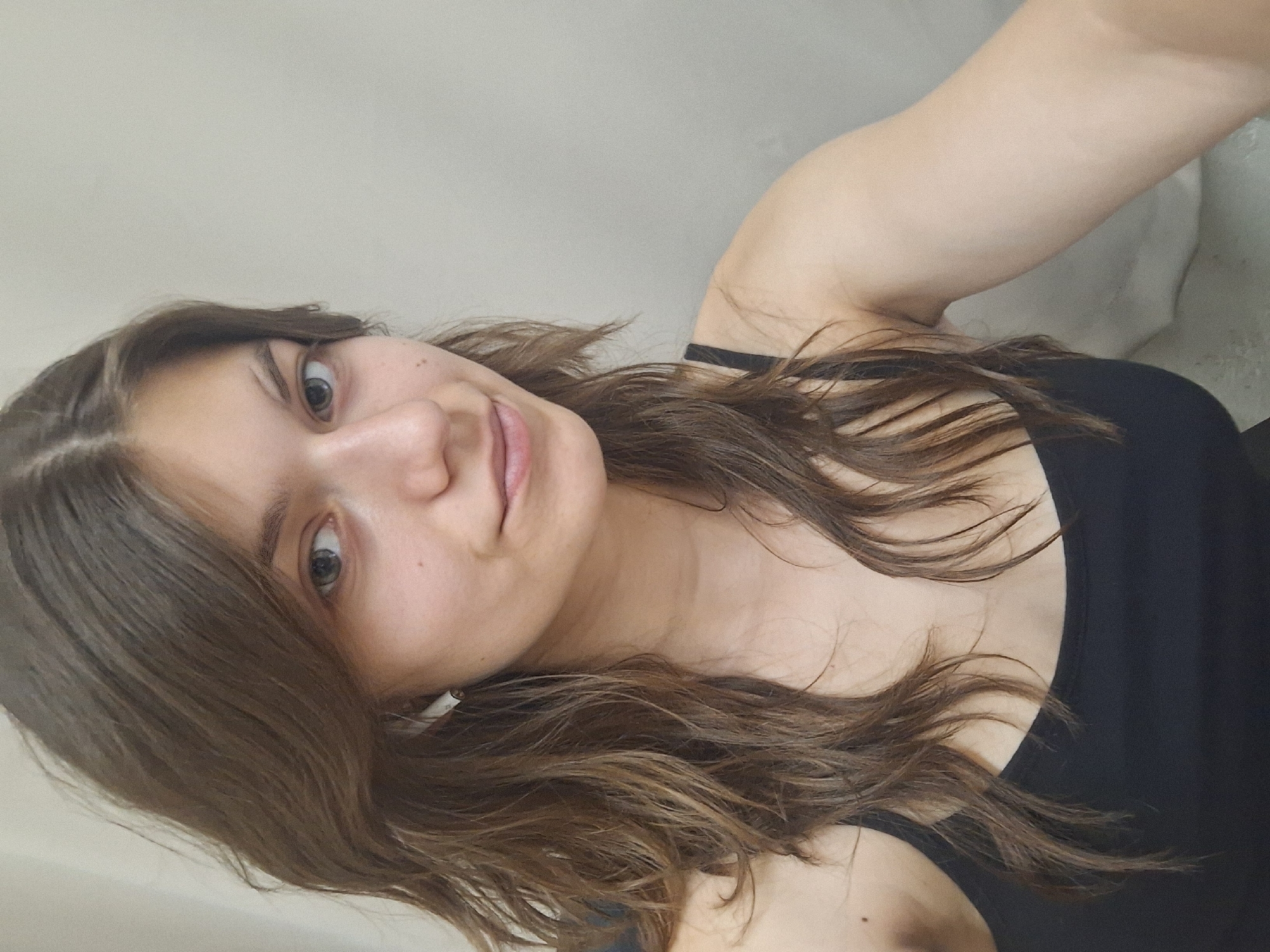 A young woman with long, brown, wavy hair is photographed from a slightly upward angle. She is wearing a black tank top. The background appears to be a plain, light-colored wall. No specific details indicate that the image was taken in Panagyurishte, Bulgaria.