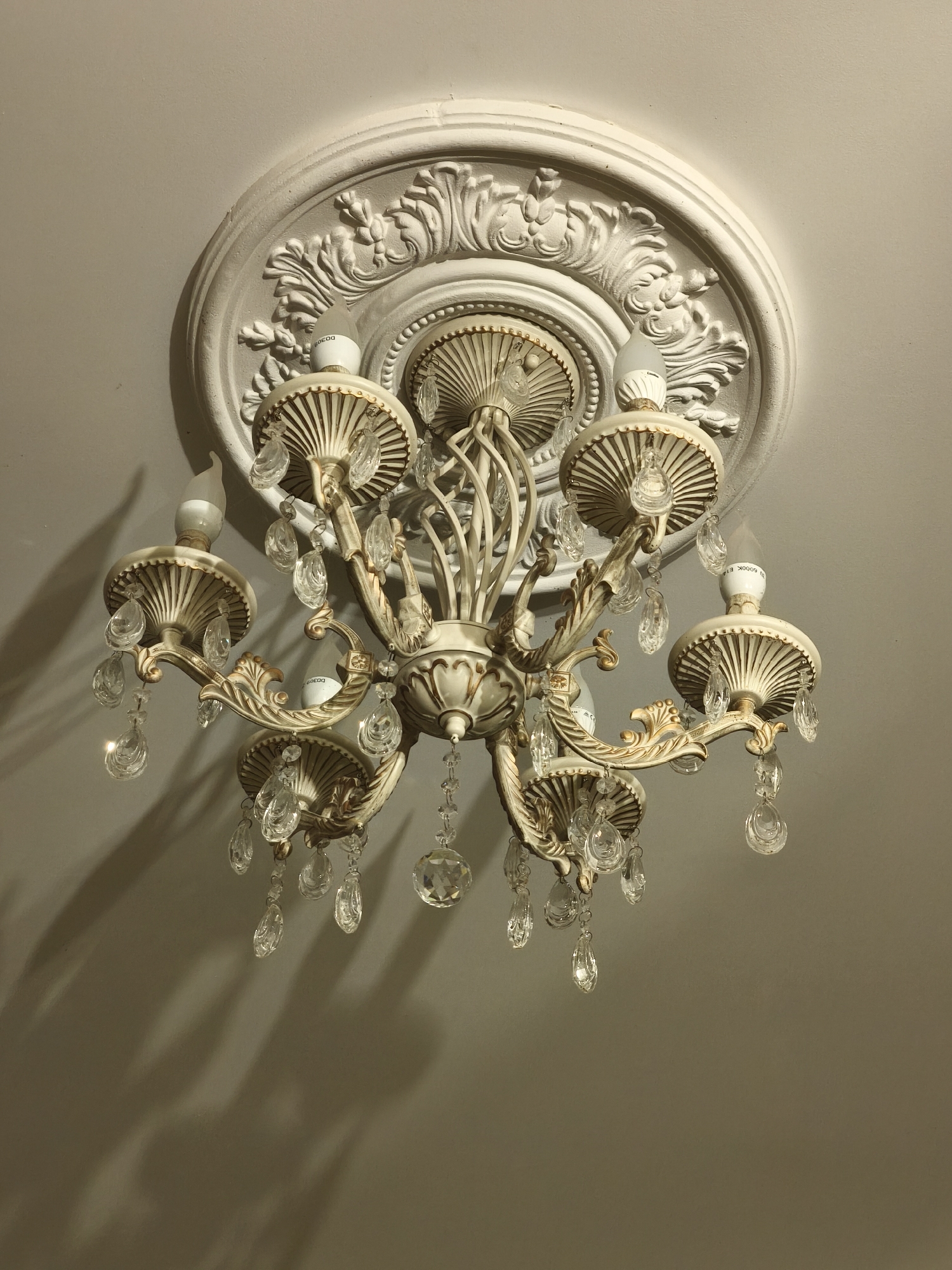 This is a close-up, low-angle, medium shot of a vintage-style chandelier hanging from a white, ornate ceiling medallion. The chandelier is made of a light-colored metal, possibly brass or a painted finish, with intricate scrollwork and ribbed shades. Several clear glass or crystal teardrop-shaped prisms are suspended from the arms of the chandelier, catching the light. The lightbulbs themselves appear to be modern LED bulbs, some of which are partially visible and labeled with Chinese characters. The ceiling is a plain, off-white surface. Shadows from the chandelier and its prisms are cast on the ceiling below, indicating a light source from above and to the right. There are no people or animals present in the image. The setting appears to be indoors, in a residential or possibly a public space with a decorative ceiling.