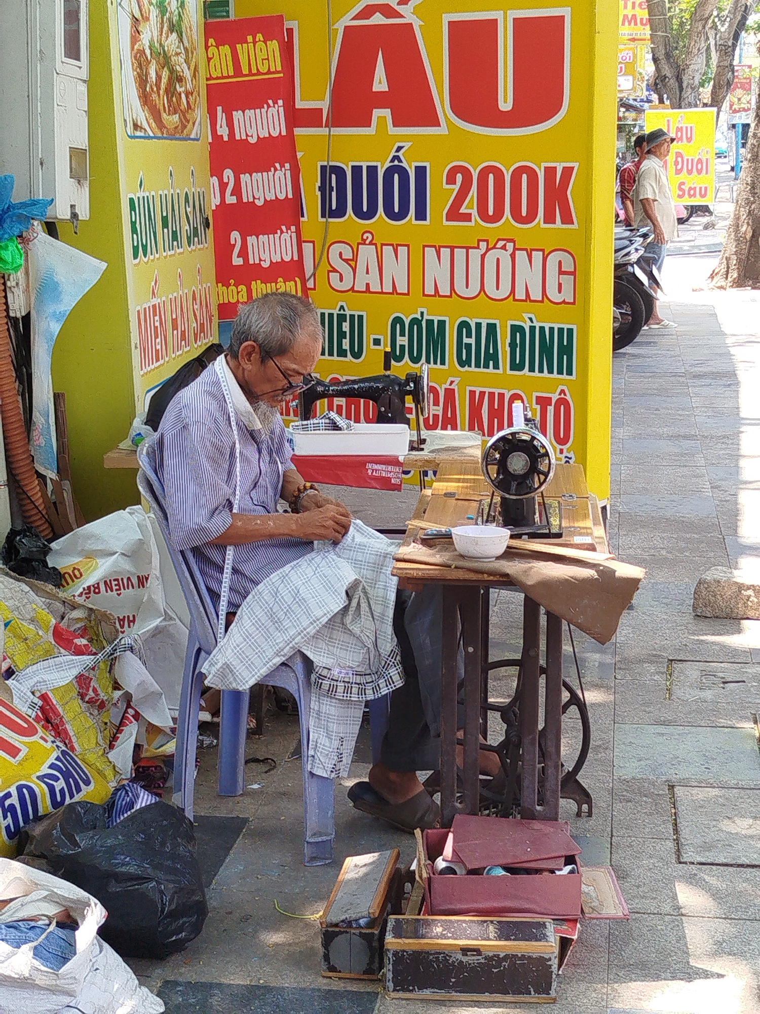 An elderly man in Ho Chi Minh City, Vietnam, is seated on a blue plastic chair, meticulously hand-sewing a plaid garment. A measuring tape is draped around his neck. He is positioned next to an antique sewing machine on a metal stand. A white bowl sits on the sewing machine table. A yellow food stall sign is prominently displayed behind him. A second sign behind the food stall sign is red and advertises deals. In the background, pedestrians and motorbikes move along the sidewalk. Various bags and boxes, including a box of sewing supplies, surround the man on the ground.