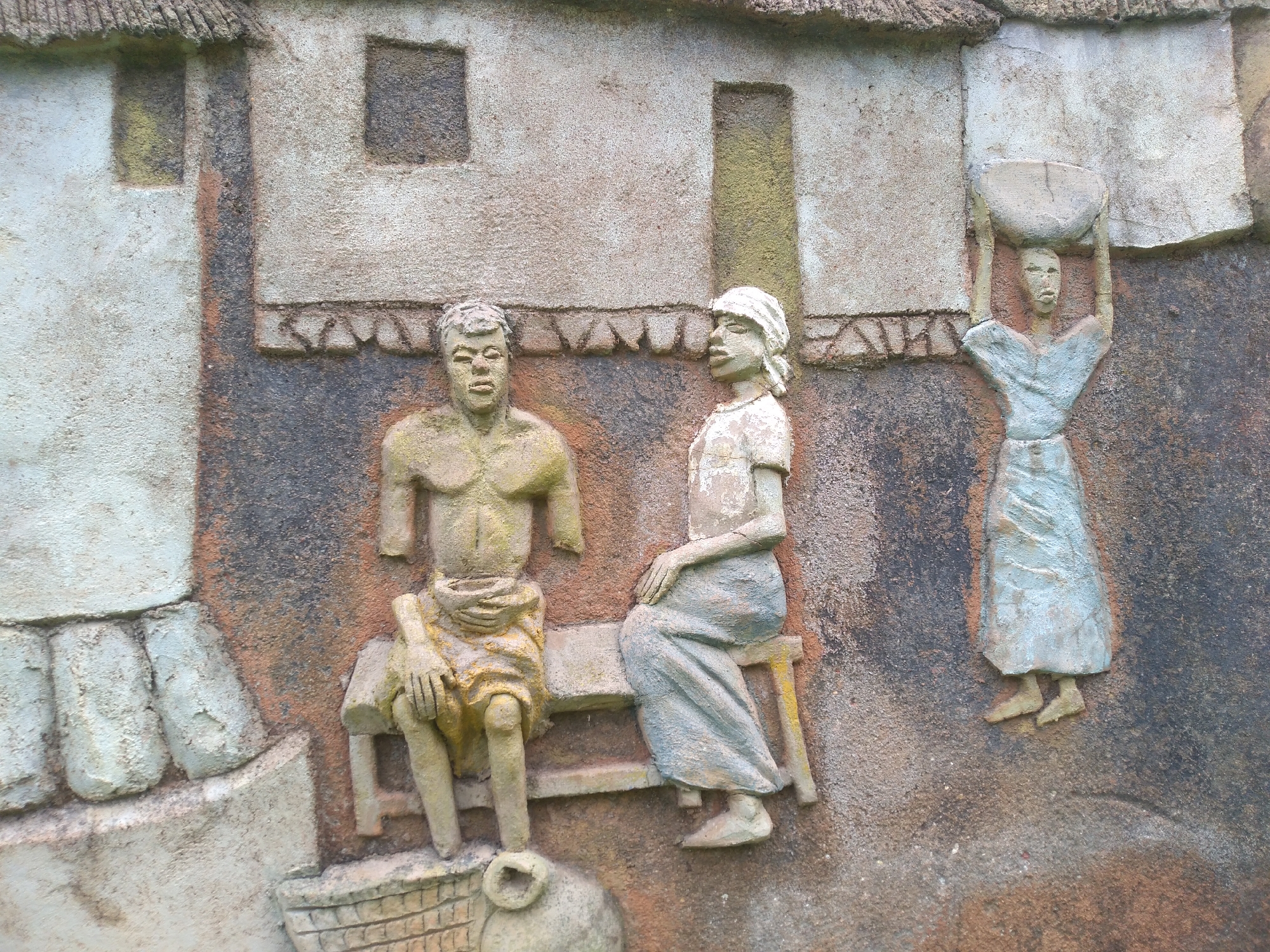 This is a bas-relief sculpture depicting a scene with three human figures set against a textured, earthy-toned wall. The sculpture appears to be located in a public space, possibly an outdoor art installation or a decorative facade in the city of Jos, Nigeria.
On the left, a muscular male figure is seated on a bench. He is depicted with his chest exposed and wears a garment around his waist and upper legs. His hands are resting on his knees.
In the center, another figure, likely a woman, is seated on the same bench to the right of the male figure. She wears a head covering and a long, draped garment. Her hands are crossed on her lap.
To the right, a third figure, also likely a woman, stands with her arms raised, balancing a bowl or pot on her head. She is dressed in a long, flowing garment.
The background of the sculpture mimics architectural elements, with recessed rectangular shapes that suggest windows or openings, and textured surfaces resembling rough walls and roofing. The overall color palette is muted, with earthy browns, grays, and hints of green and yellow, consistent with the natural materials and climate of the region. The sculpture's detailing suggests a focus on everyday life and traditional activities.
