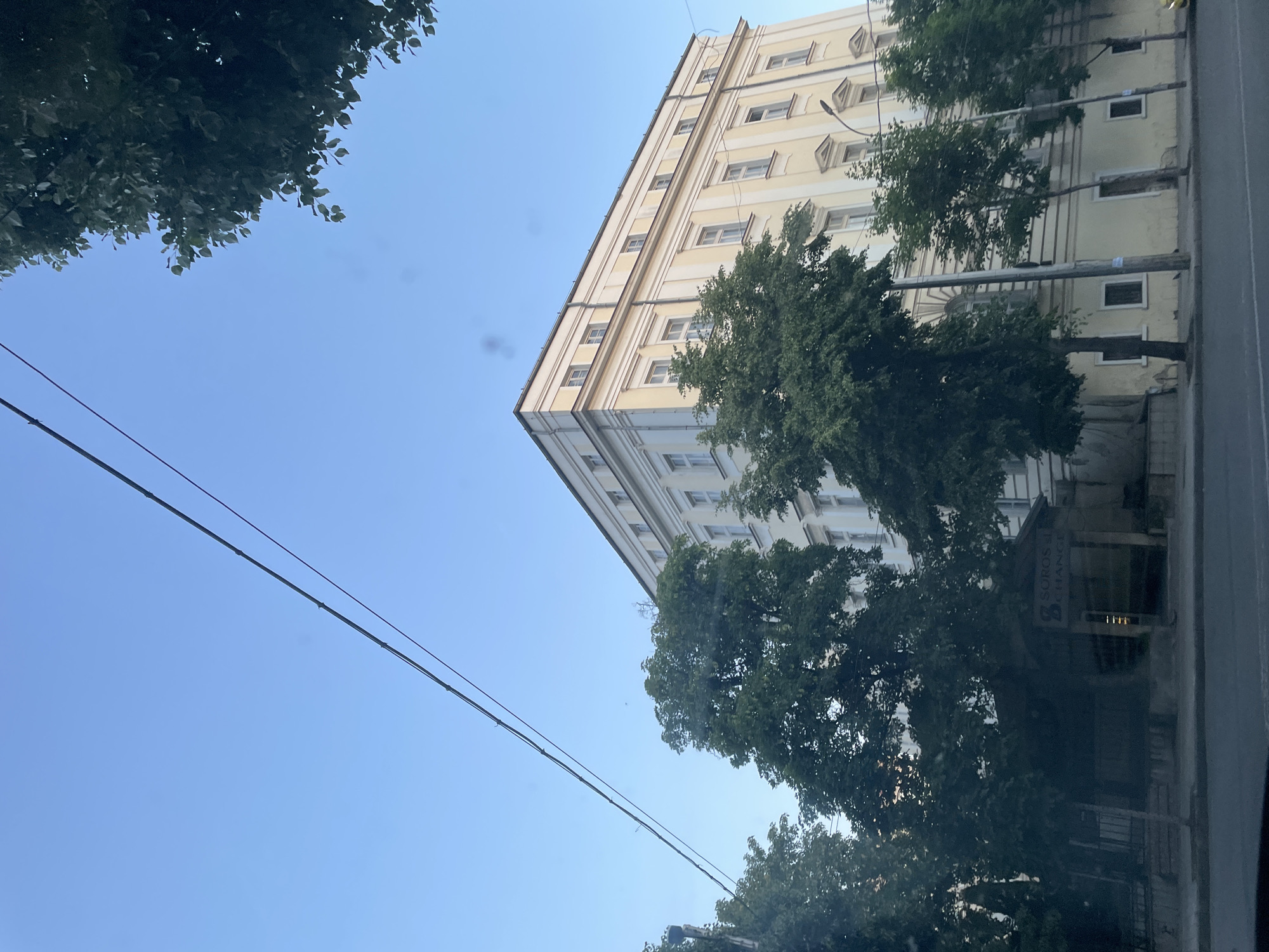 The image is a low-angle shot of a tall, light-yellow building with multiple windows in Sofia, Bulgaria. Trees partially obscure the building. Power lines stretch across a clear blue sky. The building appears to be a government building of some sort. The street level is partly visible with storefronts or offices. The environment suggests an urban setting on a sunny day. The signage "Soros de Crianza" is partially visible on one of the storefronts.