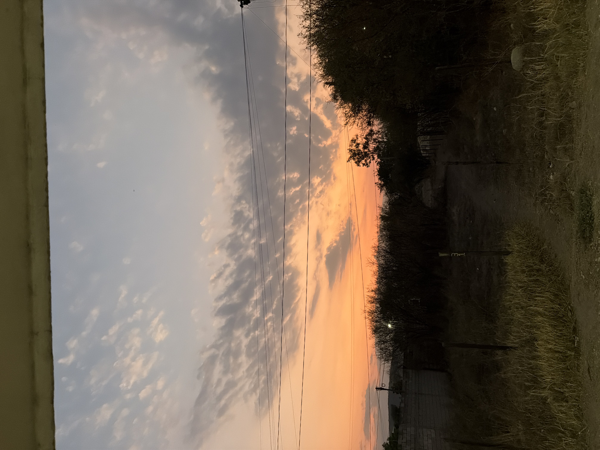 The image depicts a sunset view in what appears to be a semi-urban or rural area in Cuautla, Mexico. The sky displays a gradient of colors, ranging from a pale blue with scattered cirrus clouds at the top to a vibrant orange near the horizon, indicating the setting sun. Power lines stretch across the sky. The right side of the image features a dark, unpaved path or dirt road bordered by trees and vegetation. In the lower portion of the image, one can observe the tops of structures resembling walls and potentially houses. A concrete structure is visible on the left edge of the frame, suggesting the vantage point is from inside a building or near its exterior. The light conditions suggest it is late afternoon or early evening. No people are apparent in the image.