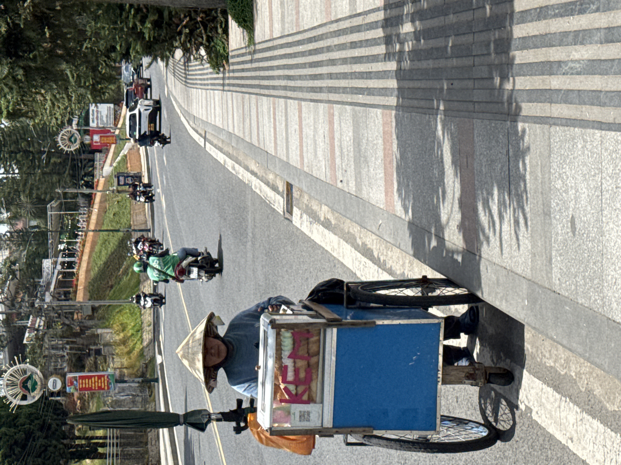 In the city of Da Lat, Vietnam, a vendor wearing a conical hat pushes a cart filled with baked goods and ice cream down a paved street. The cart has a blue base and a transparent display case filled with bread loaves and ice cream bars. The vendor is dressed in a blue long-sleeved shirt and dark pants. Behind the vendor, a person on a motorcycle in a green shirt is visible on the road, along with other motorcycles and cars. The sidewalk is paved with alternating bands of gray and pink tiles, with raised tactile paving strips. Trees and buildings are visible in the background.